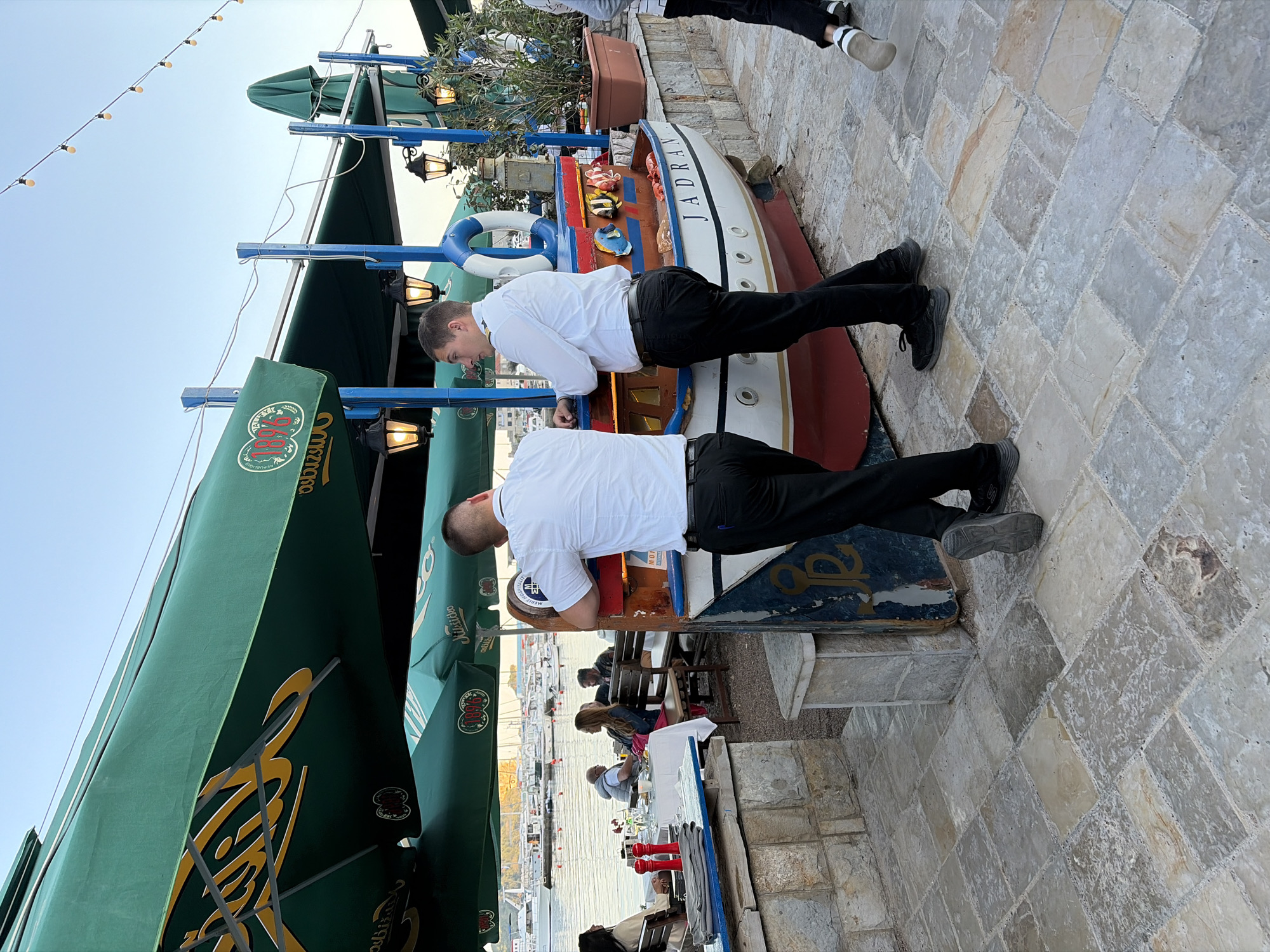 This media file captures an outdoor cafe scene by a marina, likely in Budva, Montenegro. The time of day appears to be late afternoon or early evening, with a clear sky overhead, indicated by the soft, directional light.
In the foreground, two men, presumably waiters or staff, dressed in white short-sleeved shirts and black trousers, are leaning on a decorative, boat-shaped display counter. The boat is painted white with a red hull and blue trim, and features a lifebuoy and small fish decorations. The name "JADRAN" is prominently displayed on its side in black capital letters. One man has text visible on his shirt, "MLJEVIĆ".
To the left, large green umbrellas or awnings provide shade. On one of these, the text "1894" and "Ožujsko" (a Croatian beer brand) are visible, along with other stylized lettering. String lights are strung across the top of the frame against the sky.
The ground is paved with irregularly shaped, light-colored cobblestones. In the mid-ground, other people are seated at tables, suggesting customers of the establishment. Beyond them, a body of water, likely the sea, is visible, dotted with numerous boats moored in a marina. The overall atmosphere suggests a relaxed, casual dining or leisure setting by the water.