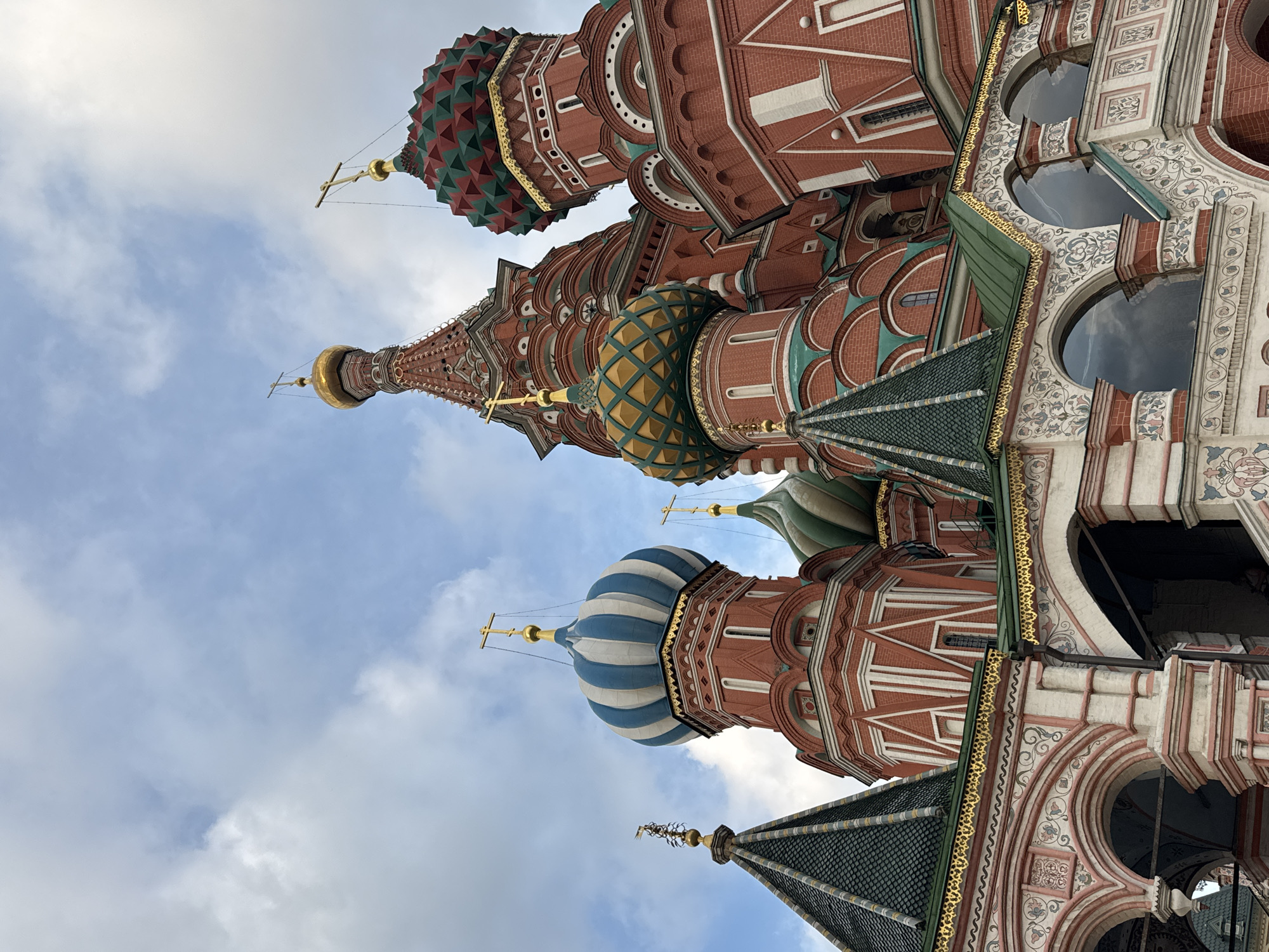 The image shows a close-up, low-angle view of the iconic Saint Basil's Cathedral in Moscow, Russia, against a backdrop of a cloudy sky. The detailed architecture of the cathedral is visible, featuring a collection of brightly colored onion domes in various patterns, including blue and white stripes, green and red diamond shapes, and gold. The walls are predominantly red brick with white trim. Several ornate crosses top the domes. The foreground showcases intricate decorative arches and upper levels of the cathedral with detailed paintings and golden borders. The scene is devoid of people or any discernible activity.