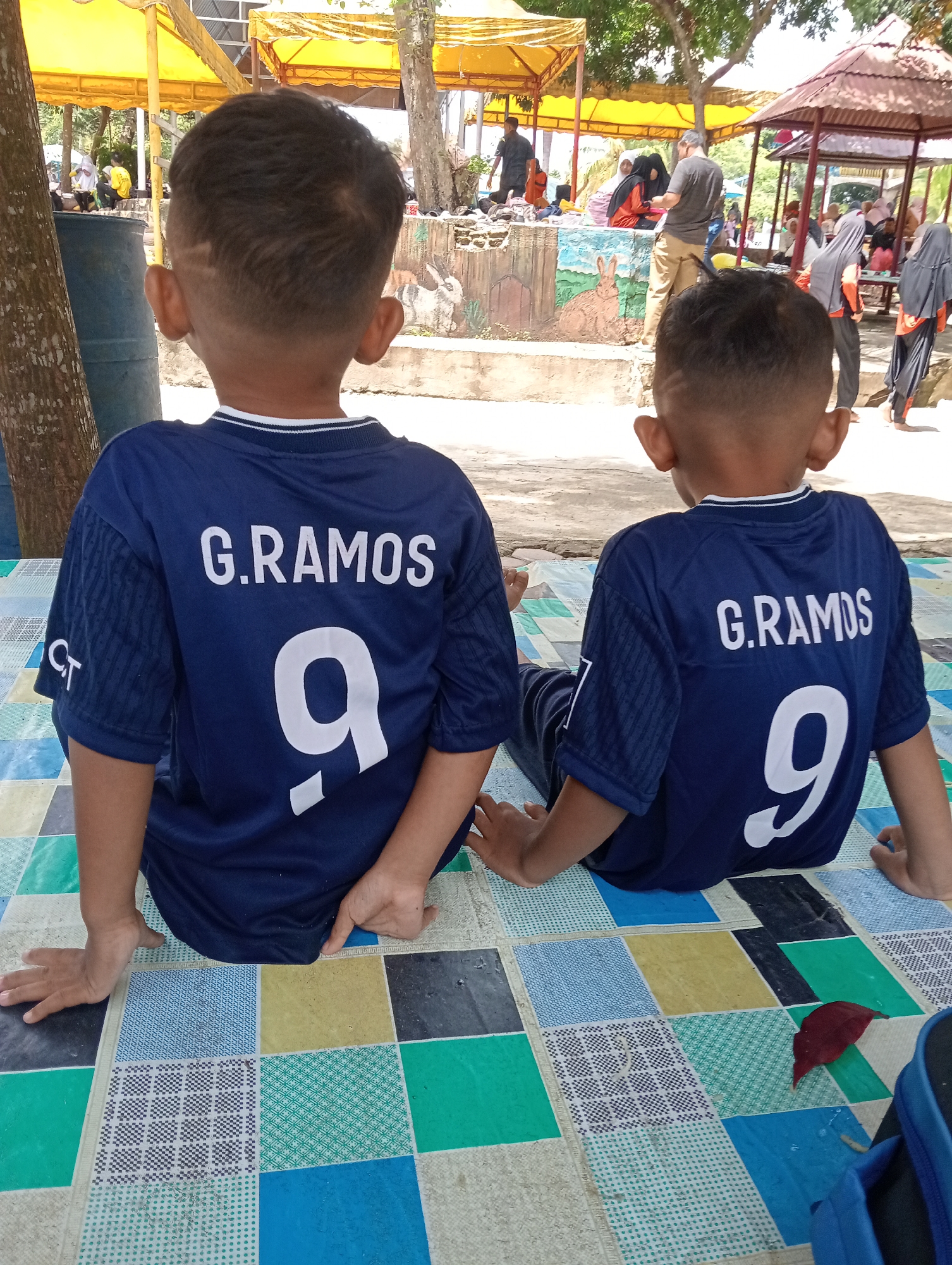 Two young boys sit side by side facing away from the camera, wearing identical navy blue soccer jerseys with "G.RAMOS" printed above the number "9" on the back. They are sitting on a multicolored tiled surface. In the background, there are several people under canopies and in a gazebo. The location is Lhokseumawe, Indonesia.