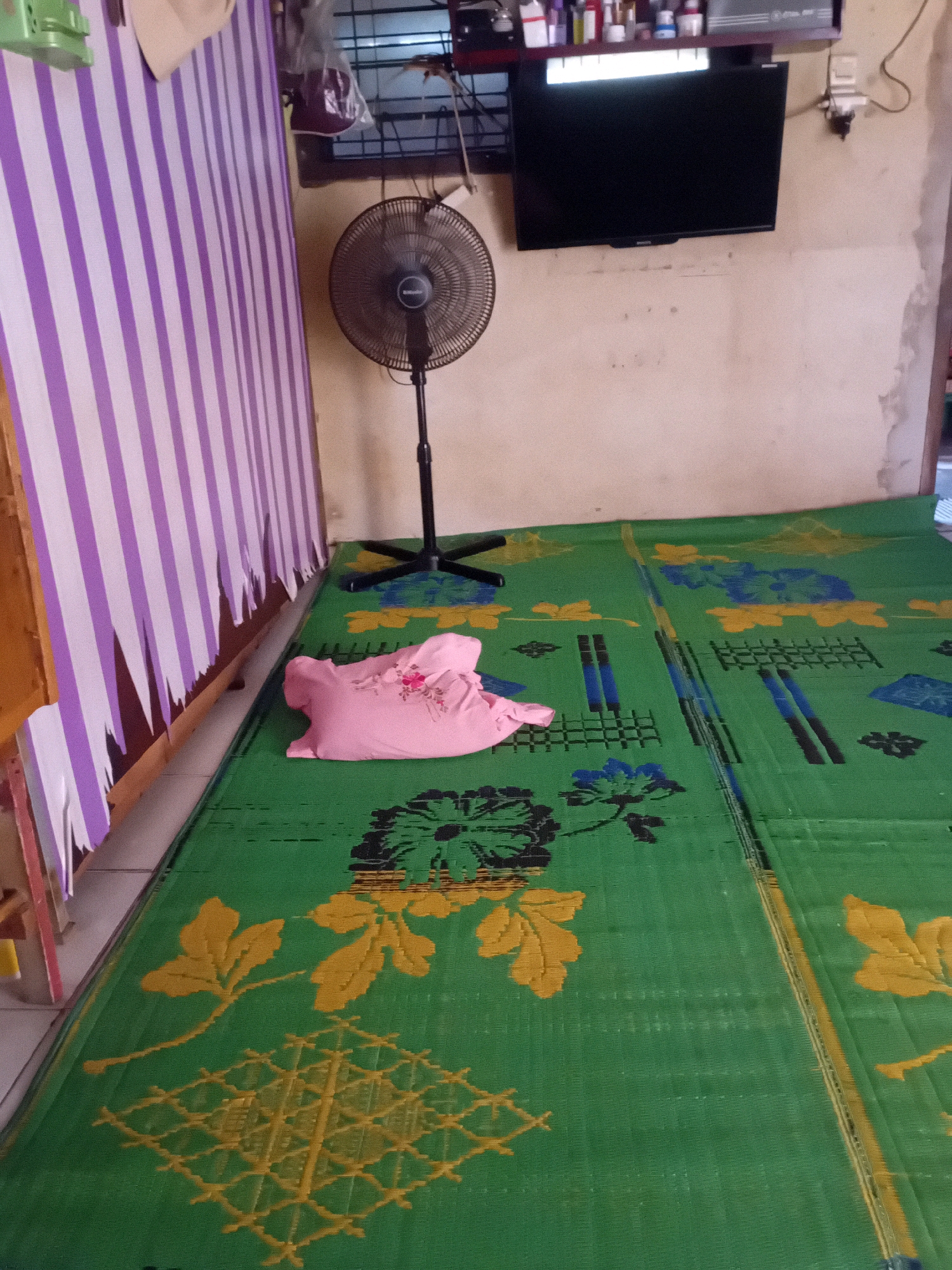 The image depicts an interior scene, likely a bedroom, with a floor covered by a green woven mat featuring floral and geometric patterns in yellow and blue. A pink folded blanket or cloth with a floral embroidery is placed on the mat. To the left, a section of a wall is covered by a striped curtain in purple and white. In the background, a black standing fan is positioned near a wall, and a black flat-screen television is mounted on the wall above. The wall itself appears to be a light beige color with some discoloration and textured patches. The overall environment suggests a simple living space. The image is likely taken in Pekanbaru, Indonesia, given the common use of such woven mats and the general aesthetic often found in residential interiors in that region.