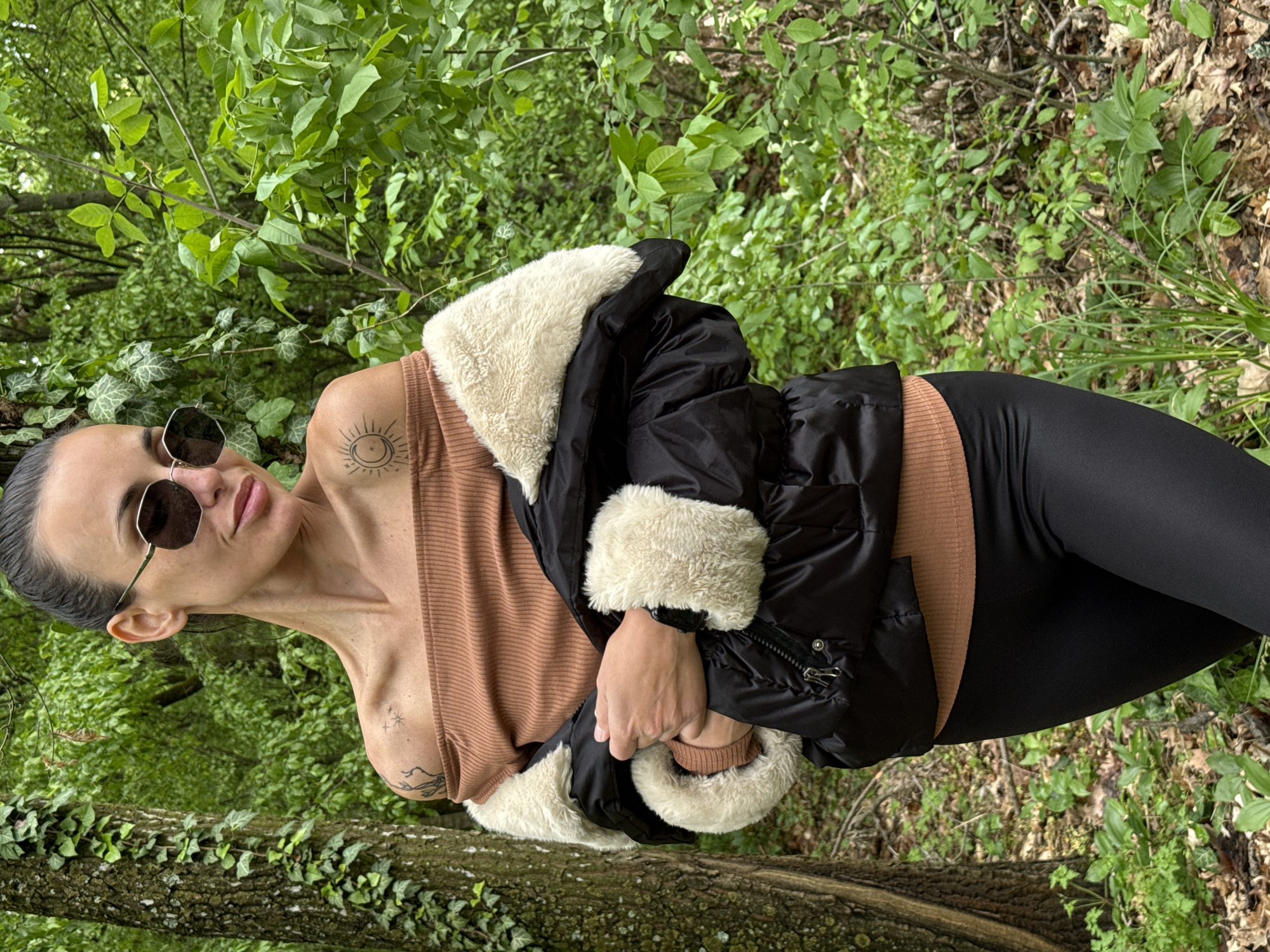 A person is lying sideways in a wooded area. The person has dark hair pulled back and is wearing sunglasses, a tan ribbed top, black leggings, and a black jacket with white fur trim. The person is resting on a tree trunk covered in green ivy and is surrounded by green vegetation. There is no indication of the city of Pleven, Bulgaria, in the image.