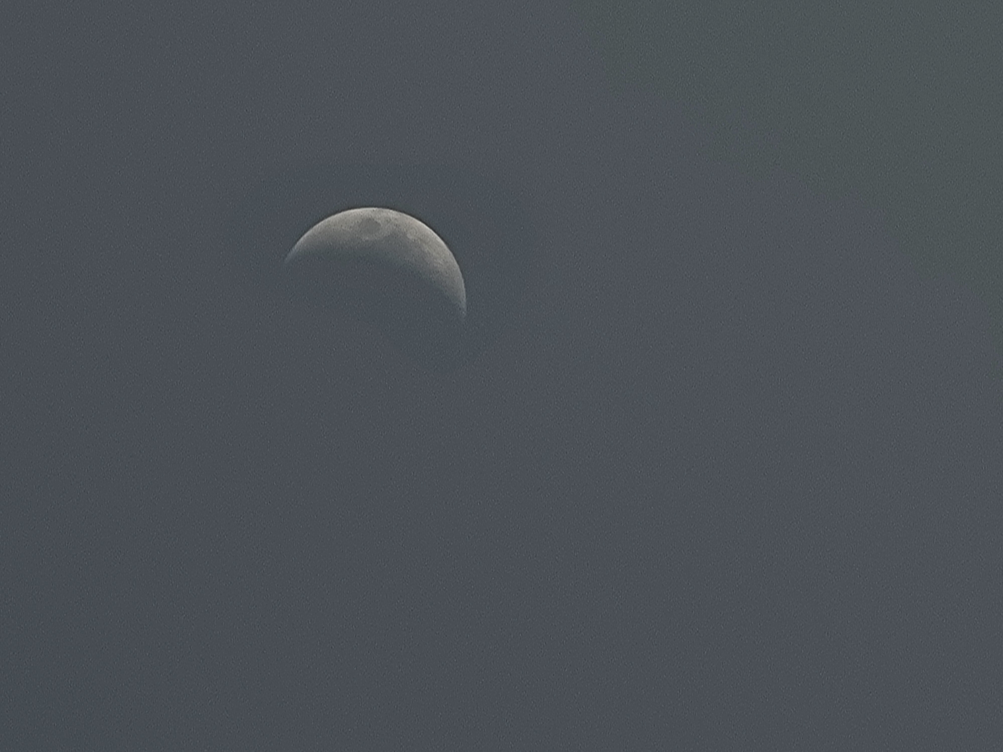 The media file shows a crescent moon in a hazy, gray sky. The moon is partially obscured by what appears to be clouds or atmospheric haze, creating a soft-focus effect. The surface of the moon is visible, with some craters and darker patches indicating lunar topography. The sky is a uniform, muted gray, suggesting an overcast or twilight condition. There are no other celestial bodies or land features visible. The overall impression is one of a serene, quiet moment in the sky. The image provides no information about people, specific objects, or ongoing activities. The time of day is likely either early morning or late evening, given the presence of the moon. There are no discernible location cues within the image itself, other than the stated context of Quarteira, Portugal. No text is visible in the media file.