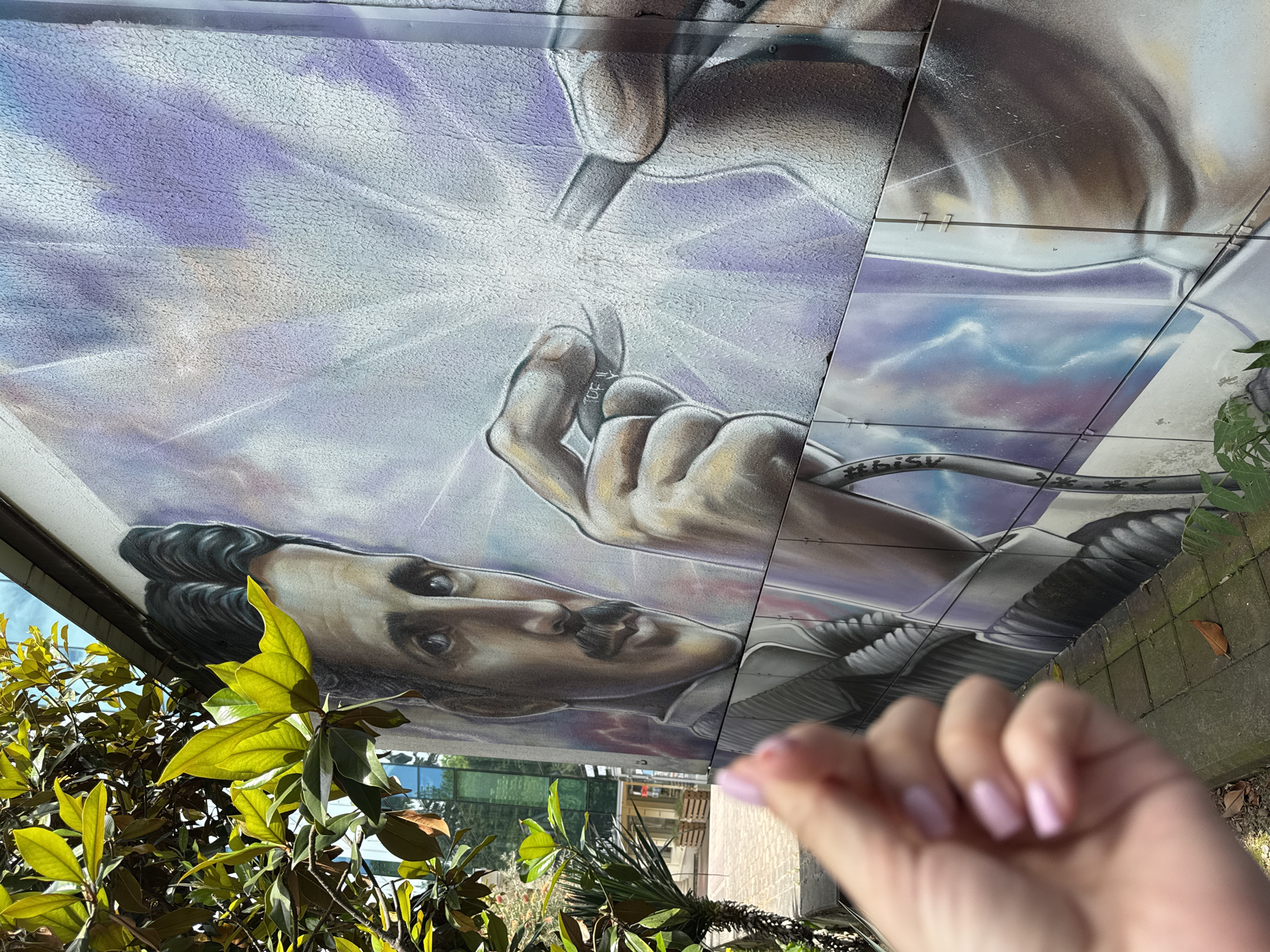 The image shows a mural of Nikola Tesla on a building in Sochi, Russia. A person's hand is in the foreground, obscuring part of the mural. The mural depicts Tesla with a mustache and dark hair, reaching out to touch a bright light emanating from two objects. The background of the mural is a swirling pattern of purple and white. A tree with green leaves is visible on the left side of the image, partially covering the mural. The building appears to be located in an urban area, with a street visible in the background.