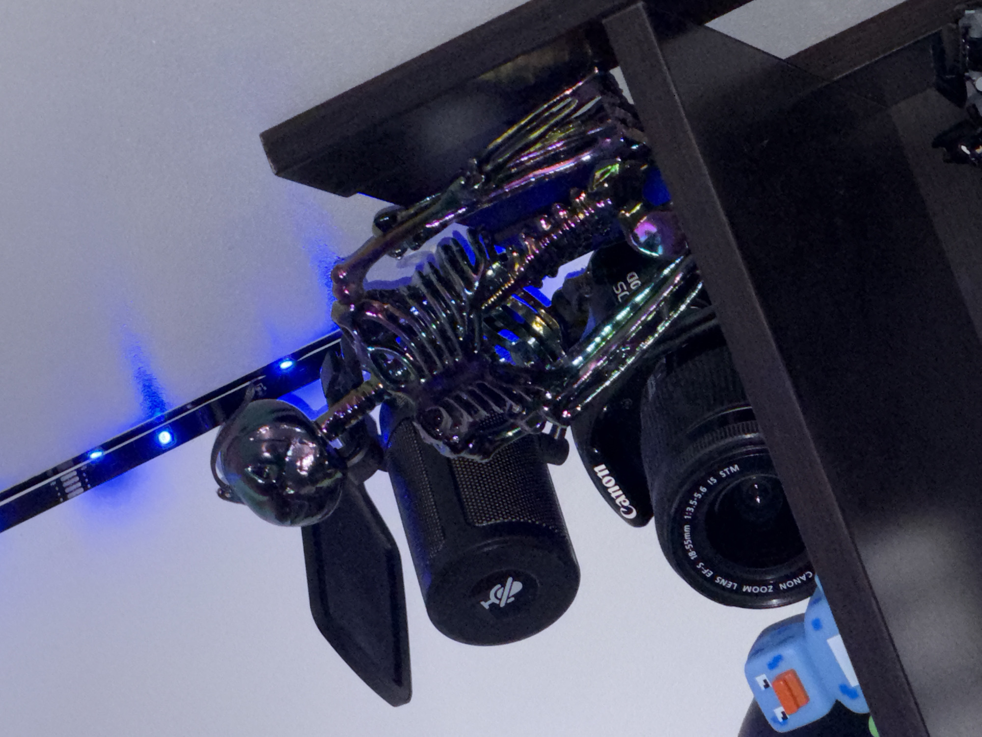 A black, iridescent skeleton figurine is positioned precariously on a dark shelf, leaning against a Canon camera lens and a black speaker. The skeleton's metallic surface reflects a spectrum of colors, enhanced by blue LED lights extending from the top left corner of the frame. A small, blue, cartoonish duck figurine is visible on the shelf to the left of the camera. The background is a plain, light-colored wall. The scene appears to be an indoor setting, likely a room or office, but there is no information to suggest its location within Oslo, Norway.