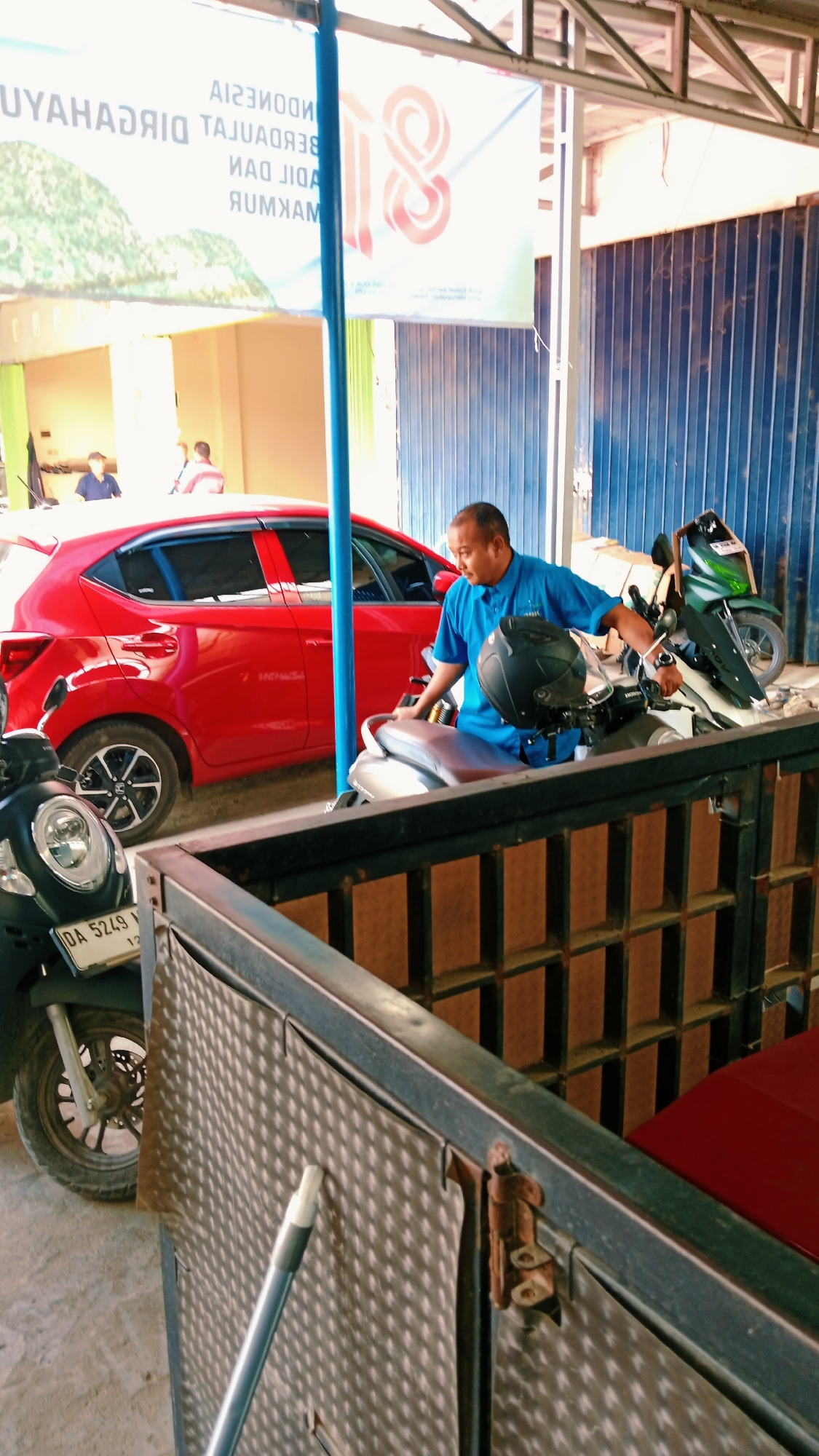 A man in a blue shirt is standing next to a motorcycle in what appears to be an open-air garage or workshop in Handil Bakti, Indonesia. A red car is parked behind him, and another motorcycle is to his left. A large banner hangs overhead with text that is partially obscured and difficult to read, but it appears to include the word "INDONESIA" and possibly "MAKMUR". The environment seems to be a commercial or industrial area, with corrugated metal walls visible in the background. The lighting suggests it is daytime, possibly late afternoon or early morning due to the shadows. The man is holding a helmet and looking down at the motorcycle.