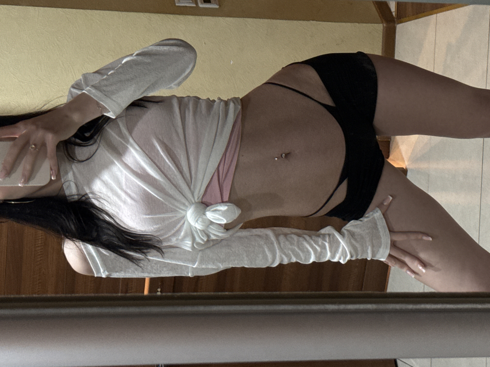 A full-body, mirror-shot photo shows a person with dark hair wearing a white, off-the-shoulder long-sleeved shirt tied at the waist with a knot, revealing a pink bralette and black high-waisted bikini-style bottoms. The person has a navel piercing and is wearing a ring on their right ring finger. The background appears to be an indoor setting, possibly a residential room, with a wooden door visible on the left and a yellow textured wall on the right. The floor is tiled with white and beige patterns. No specific details indicating the location in the city of Khimki, Russia, are present in the image.