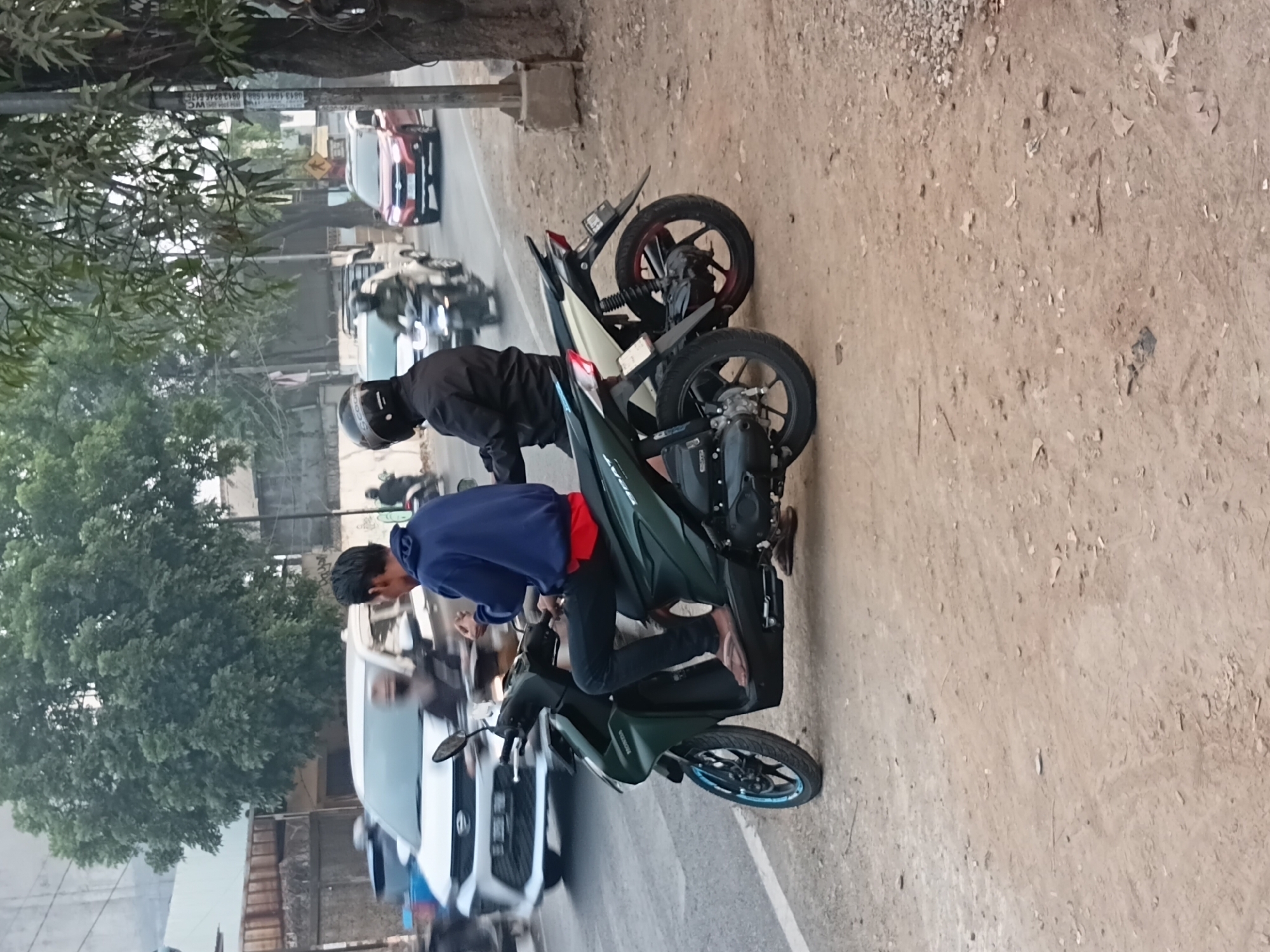 Two men are on motorcycles on a dirt road beside a paved road in Bekasi, Indonesia. The man in the foreground on a dark green motorcycle is wearing a blue hoodie, dark pants, and flip-flops. The man behind him on a white motorcycle is wearing a black jacket and helmet. In the background, other vehicles, including cars and motorcycles, are on the paved road. Trees and buildings are visible on the side of the road. The scene appears to be during the daytime.