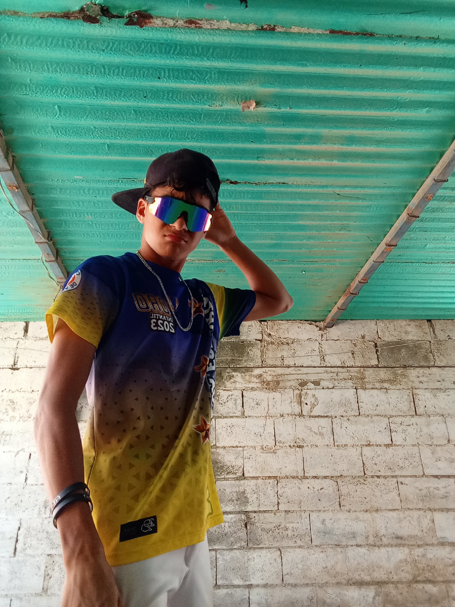 A young man is standing in front of a white brick wall under a corrugated green metal roof. He is wearing a blue and yellow shirt, white shorts, a black baseball cap, and iridescent sunglasses. He has a silver chain around his neck and black bracelets on his left wrist. He has his right hand raised to his head. The image was taken in Juan Griego, Venezuela.