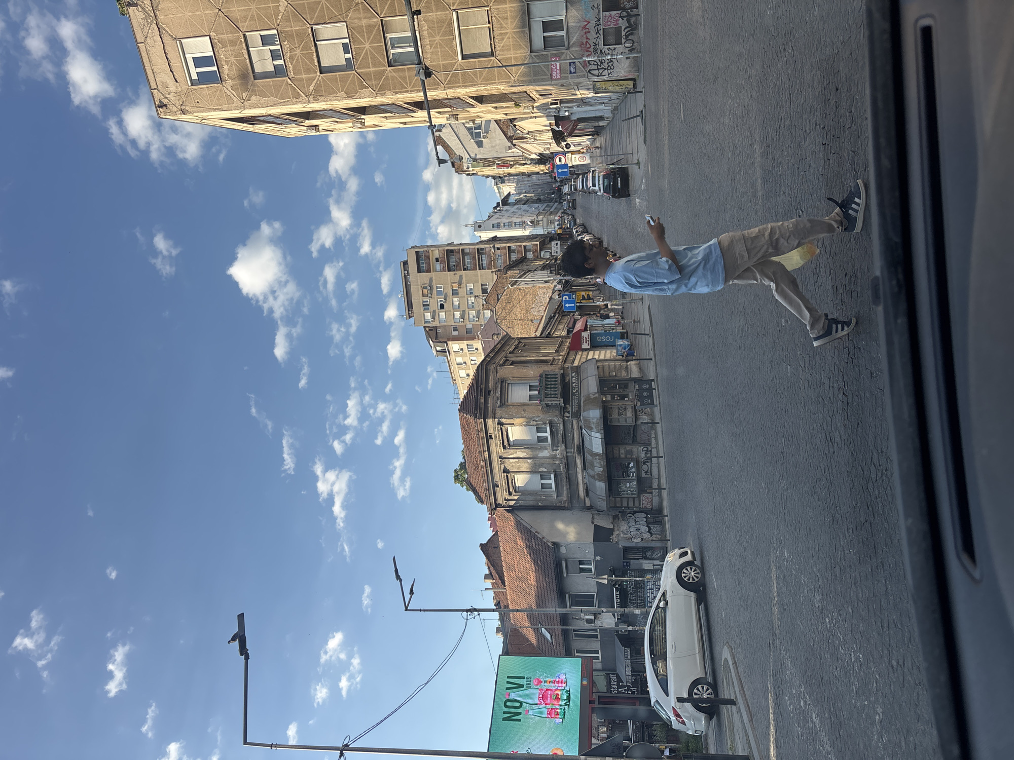 The image shows a street in Belgrade, Serbia, on a sunny day. A person is walking on the right side of the street, looking at their phone. The street is lined with buildings of varying ages and architectural styles. There is a white car parked on the side of the road. A large advertisement for NOVI water is visible on the left. The sky is blue with scattered white clouds.