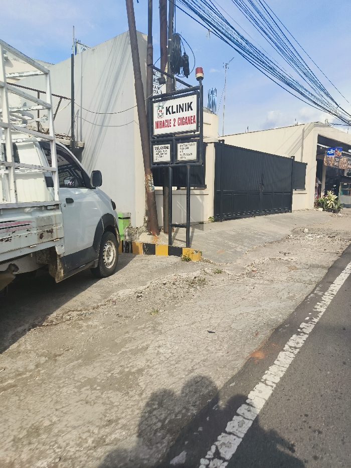 A white pickup truck is parked in front of a building with a black gate. The building has a sign that reads "Klinik Miracle 2 Ciganea." The sign is mounted on a pole with a black metal frame. The street is paved with asphalt and has a white line in the middle. The location is Jatiluhur, Indonesia. There are no people visible in the image. The image is taken from a street level perspective and appears to be taken on a sunny day.