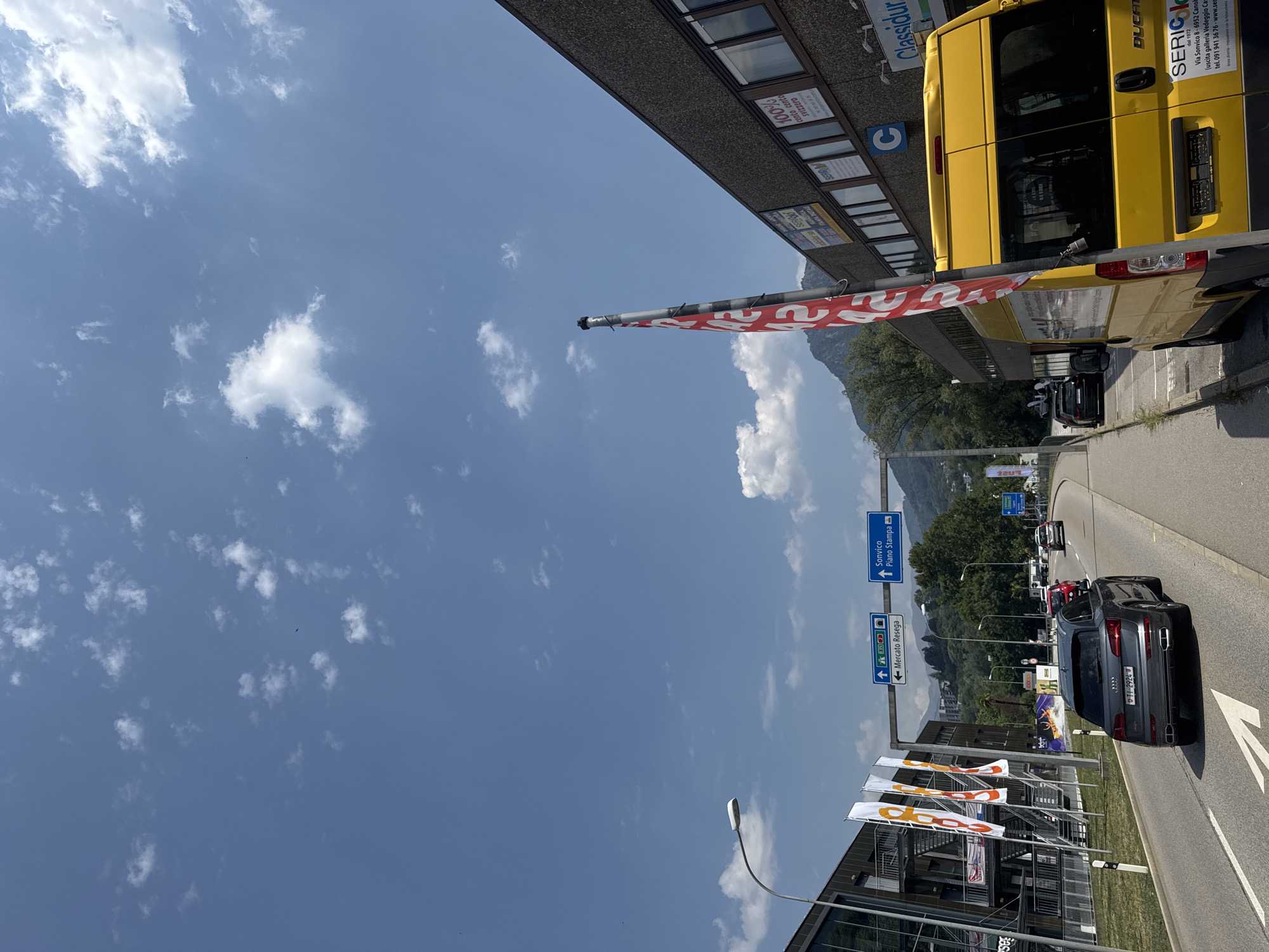 The image shows a street scene in Canobbio, Switzerland, under a clear blue sky with scattered clouds. On the right side of the road is a modern building with a textured facade, featuring multiple windows and several signs. One prominent sign reads "100% conto cento svizzero". A bright yellow van is parked in front of the building, with the "DUCATI" logo visible on its rear door. Next to it, a pole with a red and white banner is prominently displayed.
On the left side of the road, there's another modern building with a glass facade and external staircases. Several tall, white flags are positioned in front of this building. A grey Audi SUV is driving on the road, followed by other vehicles. Overhead, blue traffic signs indicate directions for "Sonvico Piano Stampa" and "Mercato Resega". A lamppost stands on the left side of the road. The surrounding area appears to be a commercial or industrial zone, with trees and a mountain range visible in the background.