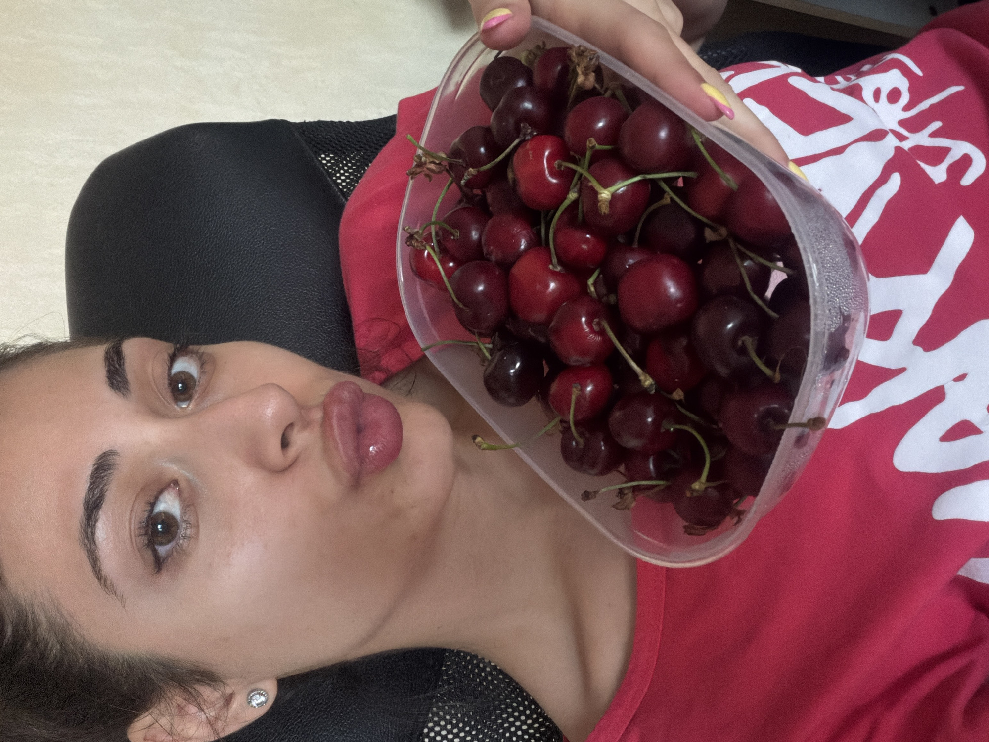 A young woman reclines in a black mesh chair, holding a clear plastic container filled with dark red cherries. She is wearing a red shirt with a white pattern. Her lips are pursed. The image appears to be taken indoors under artificial lighting. The specific location in Sofia, Bulgaria, cannot be determined from the provided image.