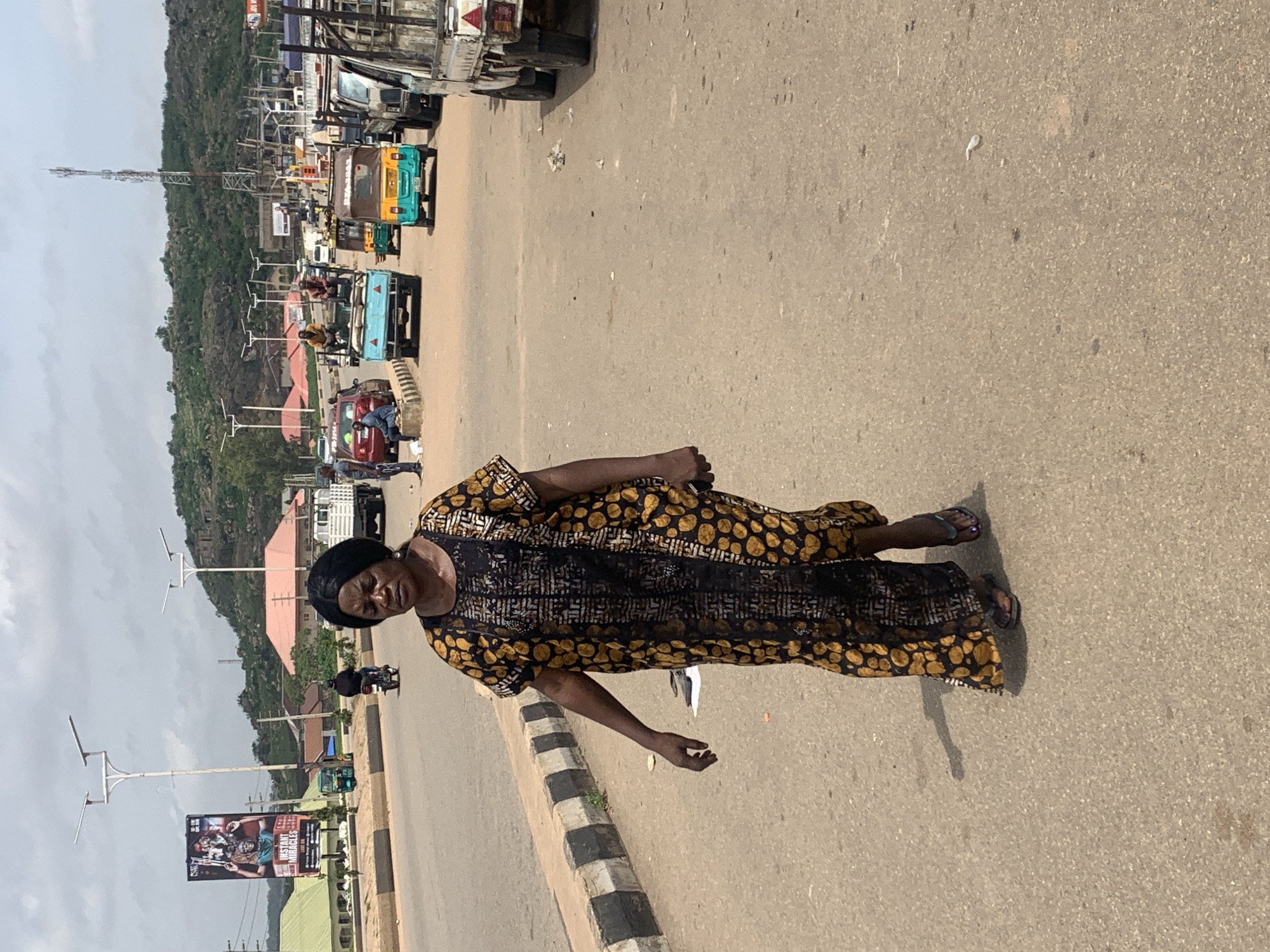 A medium shot captures a woman walking on a paved road, likely in the city of Gura Top, Nigeria, during the day. The woman, who appears to be in her 40s or 50s, is wearing a long, loose-fitting dress with a black background and a pattern of yellow circles and black and white geometric shapes. She has dark skin and dark hair styled in braids, and is wearing dark flip-flops. Her facial expression is neutral, and her gaze is directed slightly downward and to her left. She is holding a white object in her left hand, which is partially obscured by her dress.
The road is a busy thoroughfare with several vehicles visible. In the foreground, the road surface is a light brown, textured asphalt. To the left of the woman, a raised concrete curb with alternating black and white markings runs along the edge of the road. Behind her, a variety of vehicles are present, including a light blue truck with two people in the back, a yellow and green auto-rickshaw, and several other trucks and cars further in the background. A prominent billboard advertising "INSTANT MIRACLES" is visible on the left side of the frame, along with streetlights and power poles. In the distance, a hill with sparse vegetation and rocky outcrops forms the backdrop. The sky is overcast with some patches of blue. The overall impression is one of a typical urban scene in a developing country.