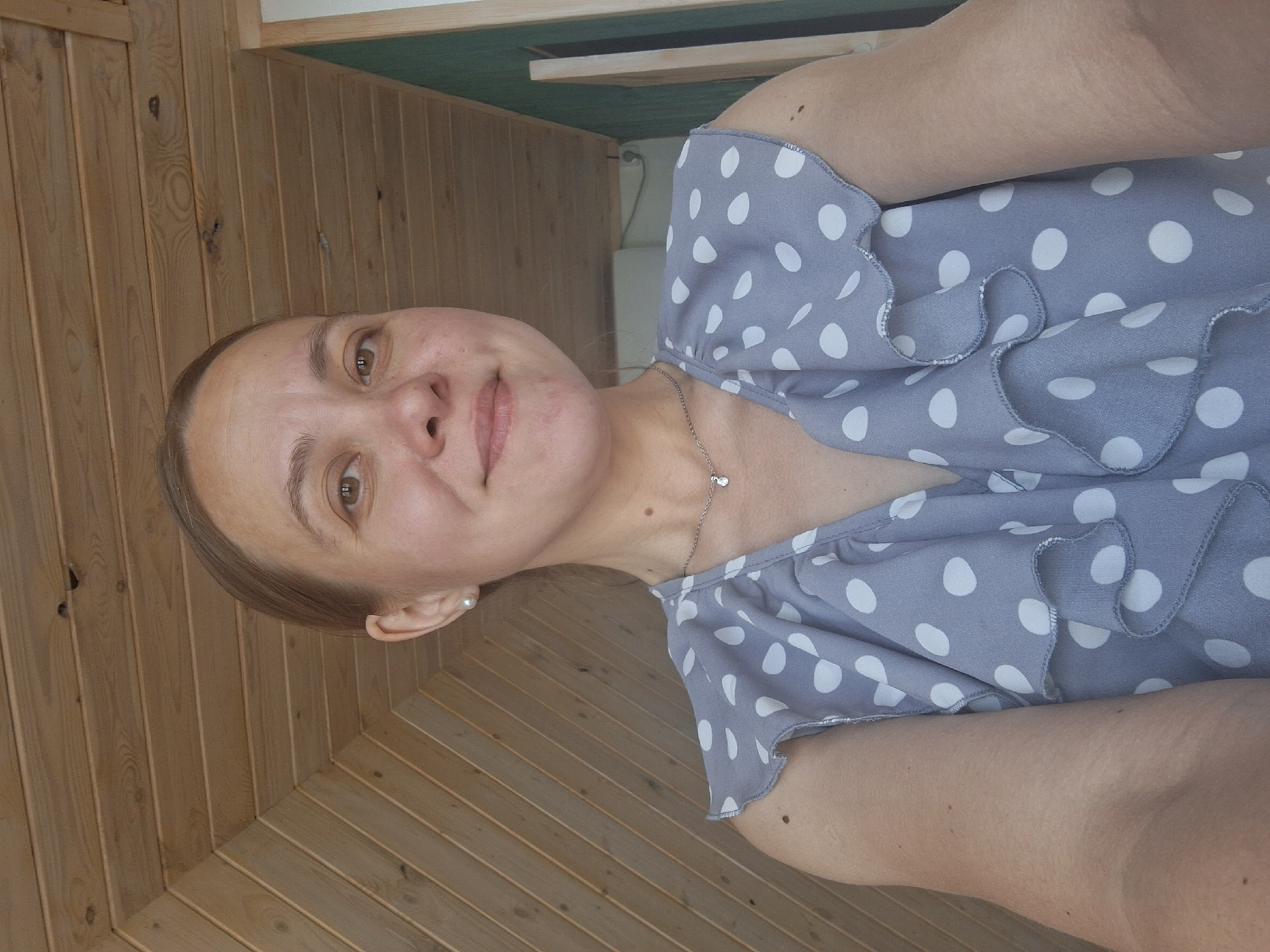 A woman with fair skin, light brown hair pulled back, and brown eyes is wearing a gray and white polka dot blouse with ruffles around the neckline and sleeves. She is also wearing a delicate silver necklace with a small pendant. She has a subtle smile. The background consists of light brown wooden paneling. The image is rotated 90 degrees clockwise. There is no verifiable information about the location of Chaikovsky, Russia, in the image.