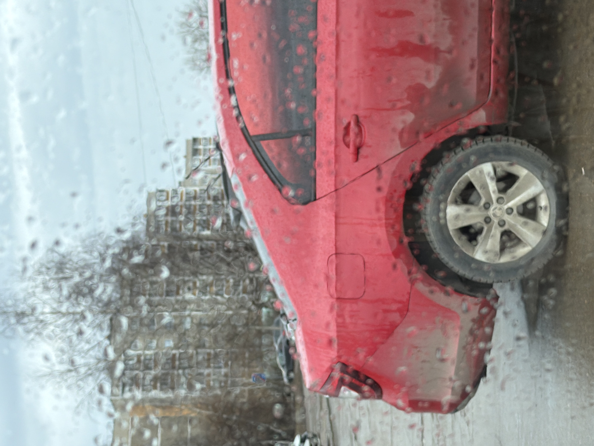 A red car is parked on the side of a street in Chaikovsky, Russia. The vehicle is dirty and has raindrops on its surface. In the background, a building and bare trees are visible through the wet glass. The image is taken from inside a vehicle during a rainy day.