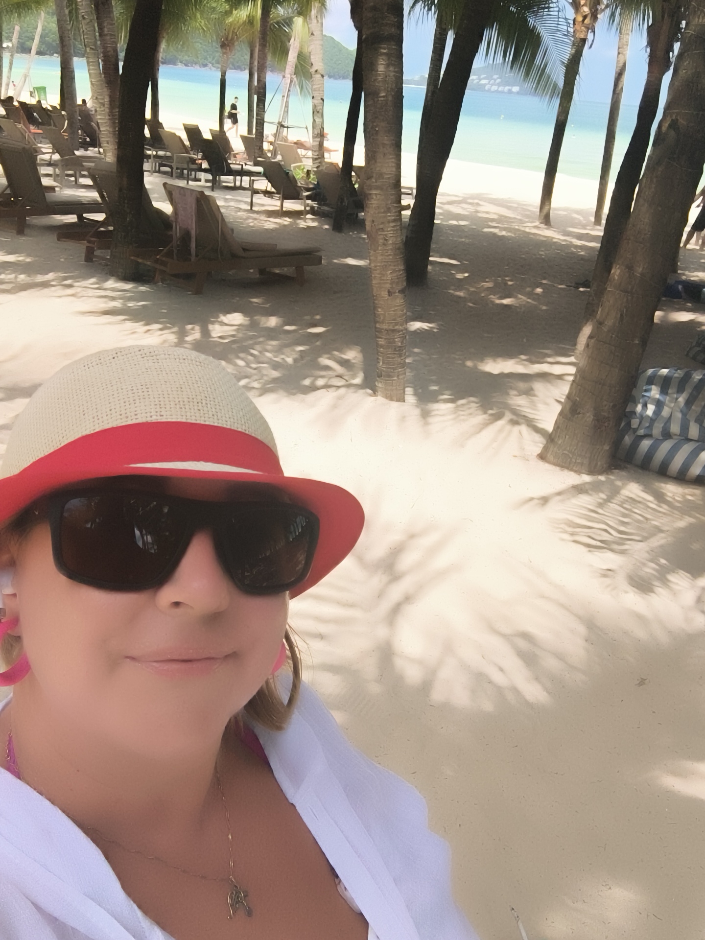 A woman wearing a straw hat with a red band and sunglasses takes a selfie on a beach in Phu Quoc, Vietnam. She is wearing a white shirt open over a pink bikini top and a necklace with a small pendant. Behind her, palm trees cast shadows on the white sand. In the background, lounge chairs are arranged on the beach, and the turquoise ocean meets the horizon.