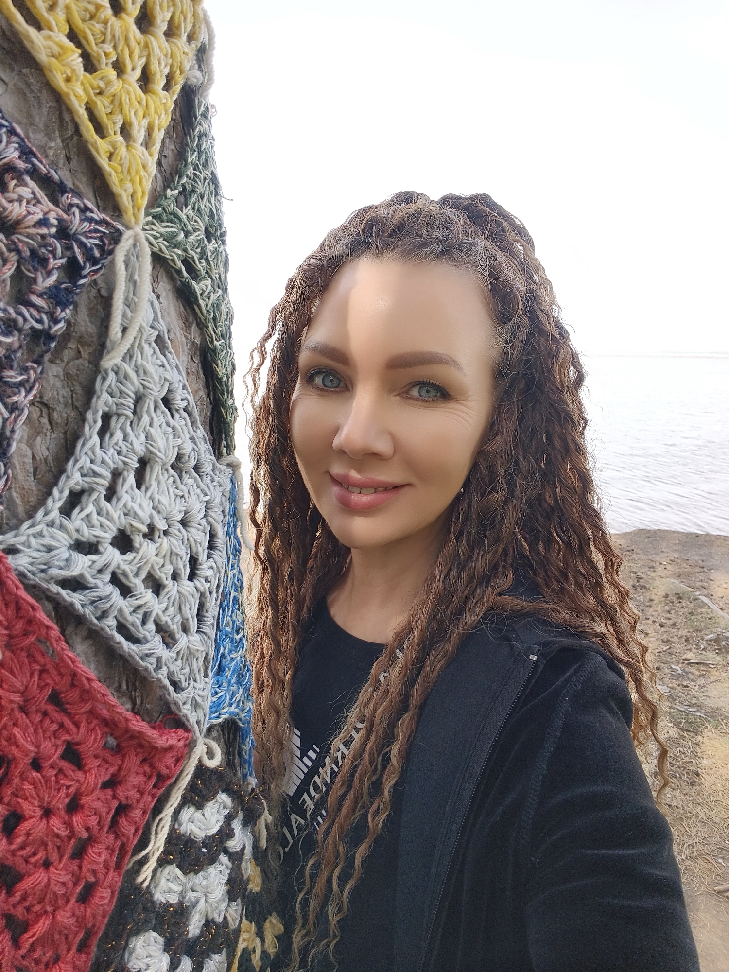 A light-skinned woman with long, brown, curly hair and blue eyes stands beside a tree adorned with colorful crocheted squares. The woman is wearing a black top and a black jacket. The background shows a body of water and a shoreline under a bright sky. The location appears to be outdoors, near the water's edge. The city and state are unknown.