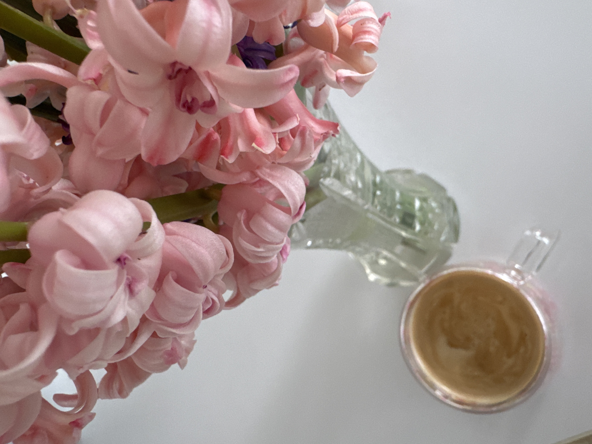 A high-angle shot presents a still life scene on a white surface. The primary focus is a bouquet of pink hyacinth flowers clustered on the left side of the frame, their dense, layered petals creating a soft, textured mass. Adjacent to the flowers, slightly blurred in the midground, is a clear glass vase. To the right, closer to the foreground, is a clear glass mug filled with a brown liquid, possibly coffee or tea. There is no indication of the location being Varna, Bulgaria. No people are visible. The environment consists of a plain white surface providing a clean backdrop.