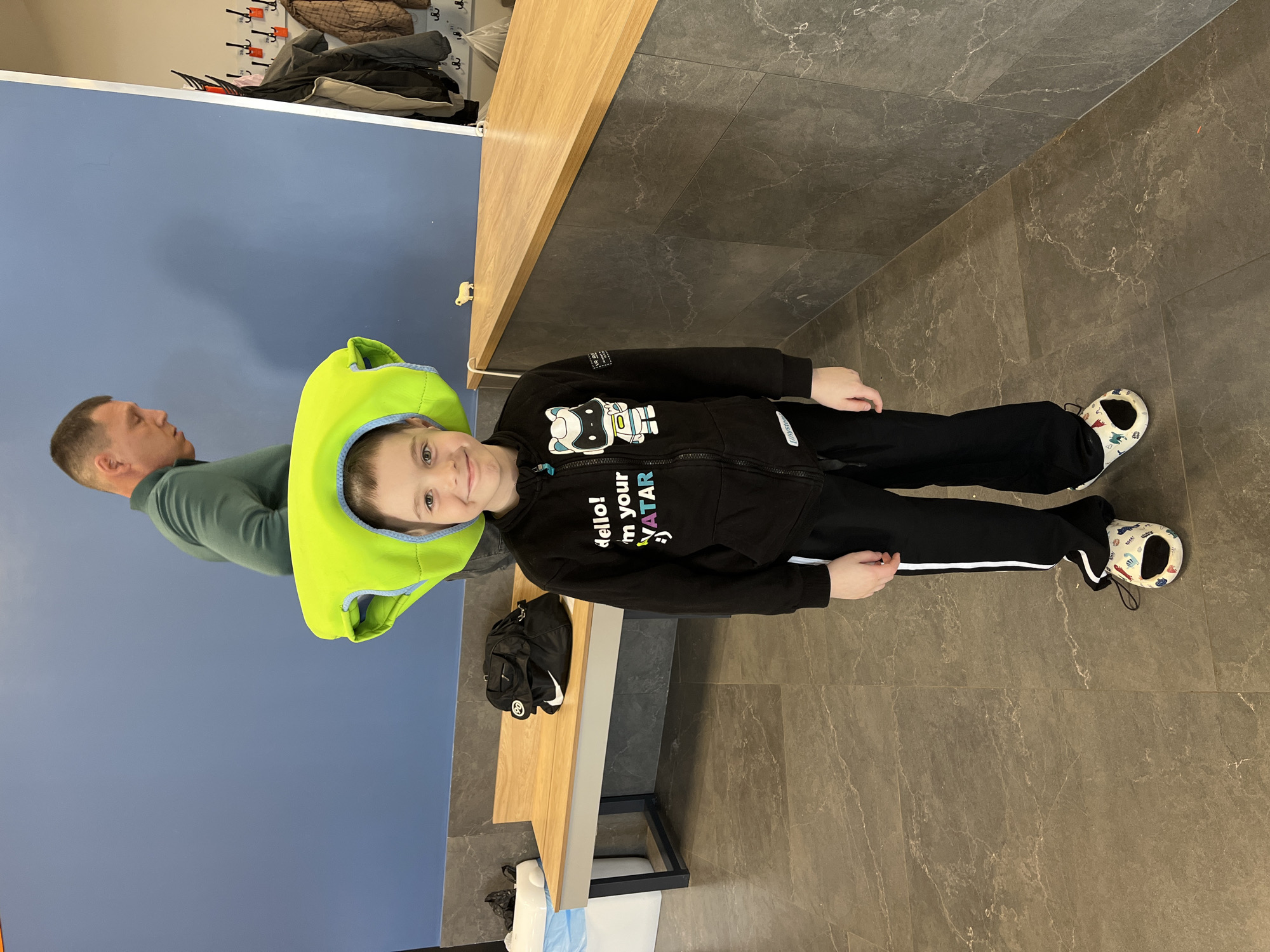 A man and a child stand next to a wall, likely indoors. The child is in the foreground, wearing black sweatpants with a white stripe, a black hoodie with lettering, and patterned shoes. The man, situated behind the child, is wearing a green shirt. The child's head is framed by a lime green life vest. They are standing on a dark tile floor. A wooden bench sits against a blue wall, with various coats and bags hung on coat hooks above. The overall setting has the feel of a public space, such as a recreational facility. Given the location (Votkinsk, Russia), it's plausible this image depicts an area within a community center, sports complex, or similar public building.