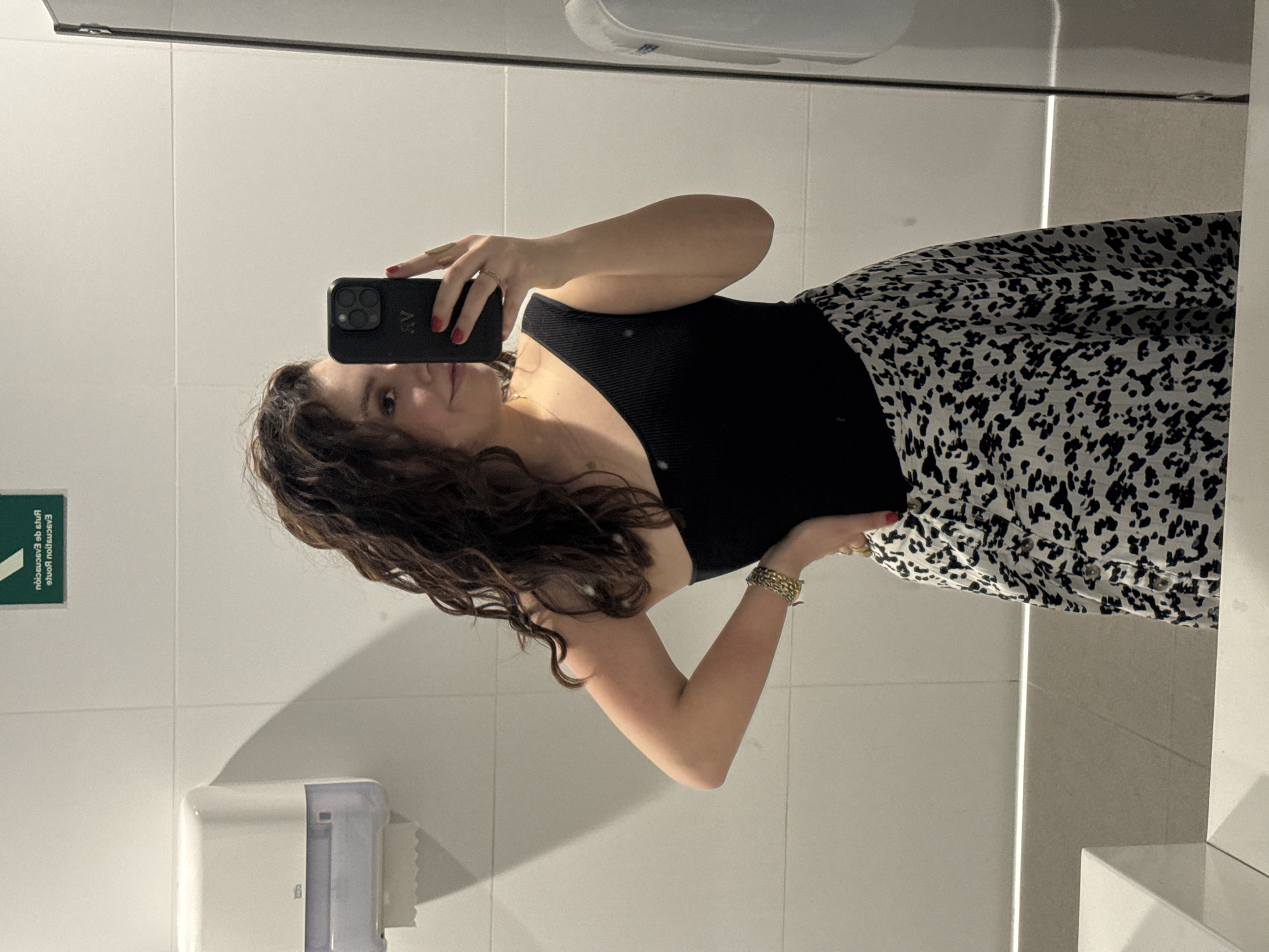 A woman is taking a selfie in a restroom. She is wearing a black top and a black and white patterned skirt. She is holding a black smartphone. The restroom has white tiled walls, a sink, and a paper towel dispenser. A green and white sign is visible on the wall. The location is likely in Mexico City, Mexico.