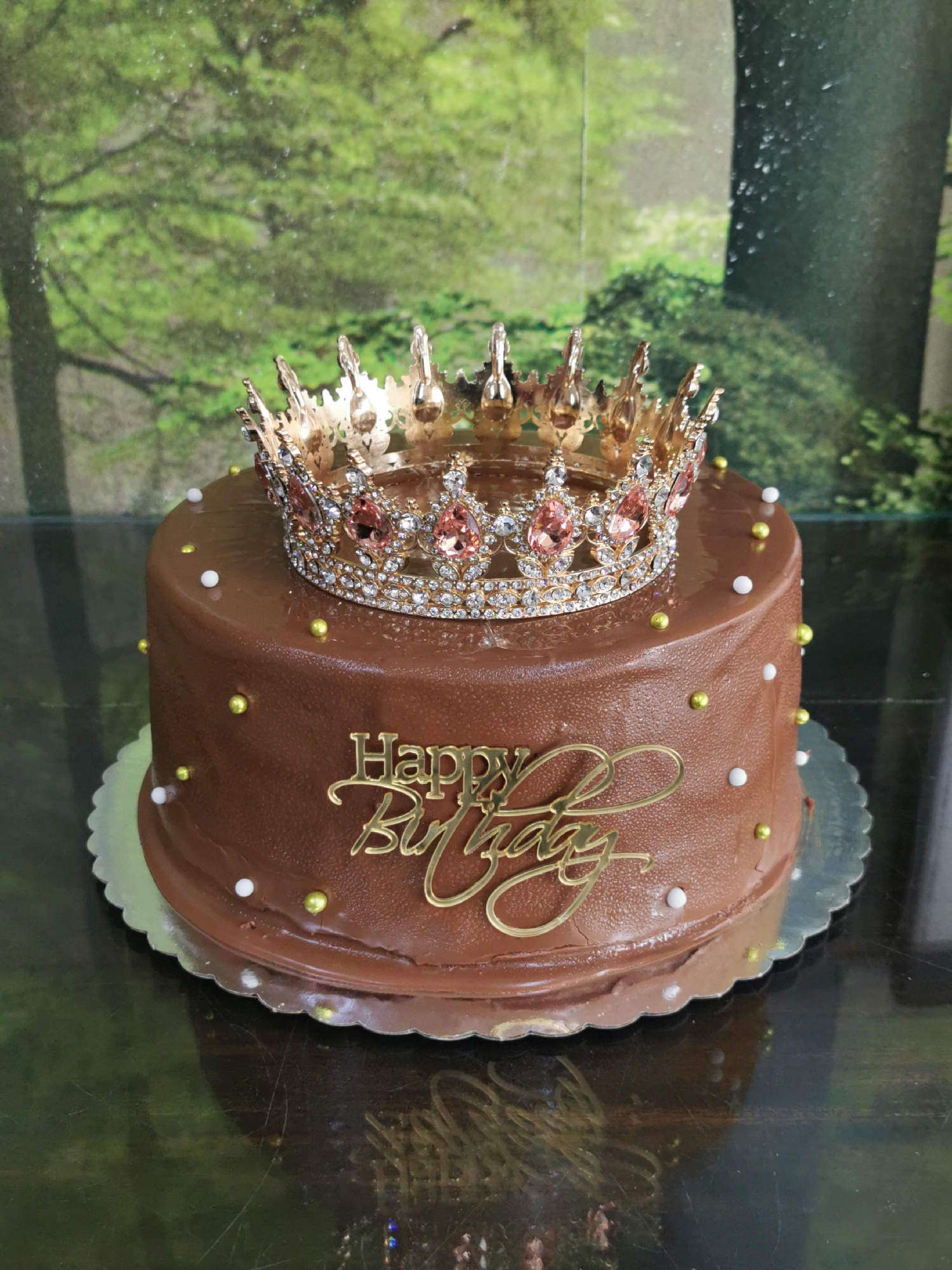 The image shows a chocolate birthday cake adorned with a gold crown and a "Happy Birthday" sign. The cake is decorated with gold and white candy pearls. The background appears to be a blurred nature scene, possibly a park or garden. The image doesn't contain enough information to identify the location as Varna, Bulgaria.