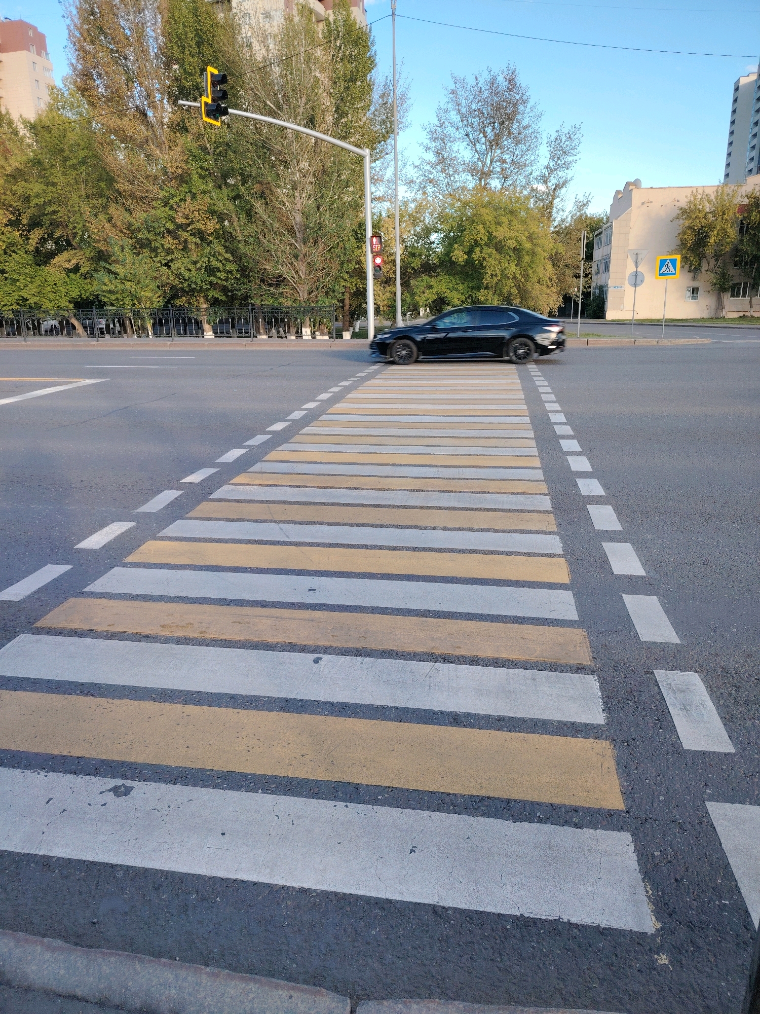 The image shows a crosswalk with alternating yellow and white stripes on an asphalt road. A black car is driving across the crosswalk, and there are traffic lights and trees in the background. The scene appears to be in a city, with buildings and a clear blue sky visible. Based on the provided information, this image was taken in Astana, Kazakhstan.