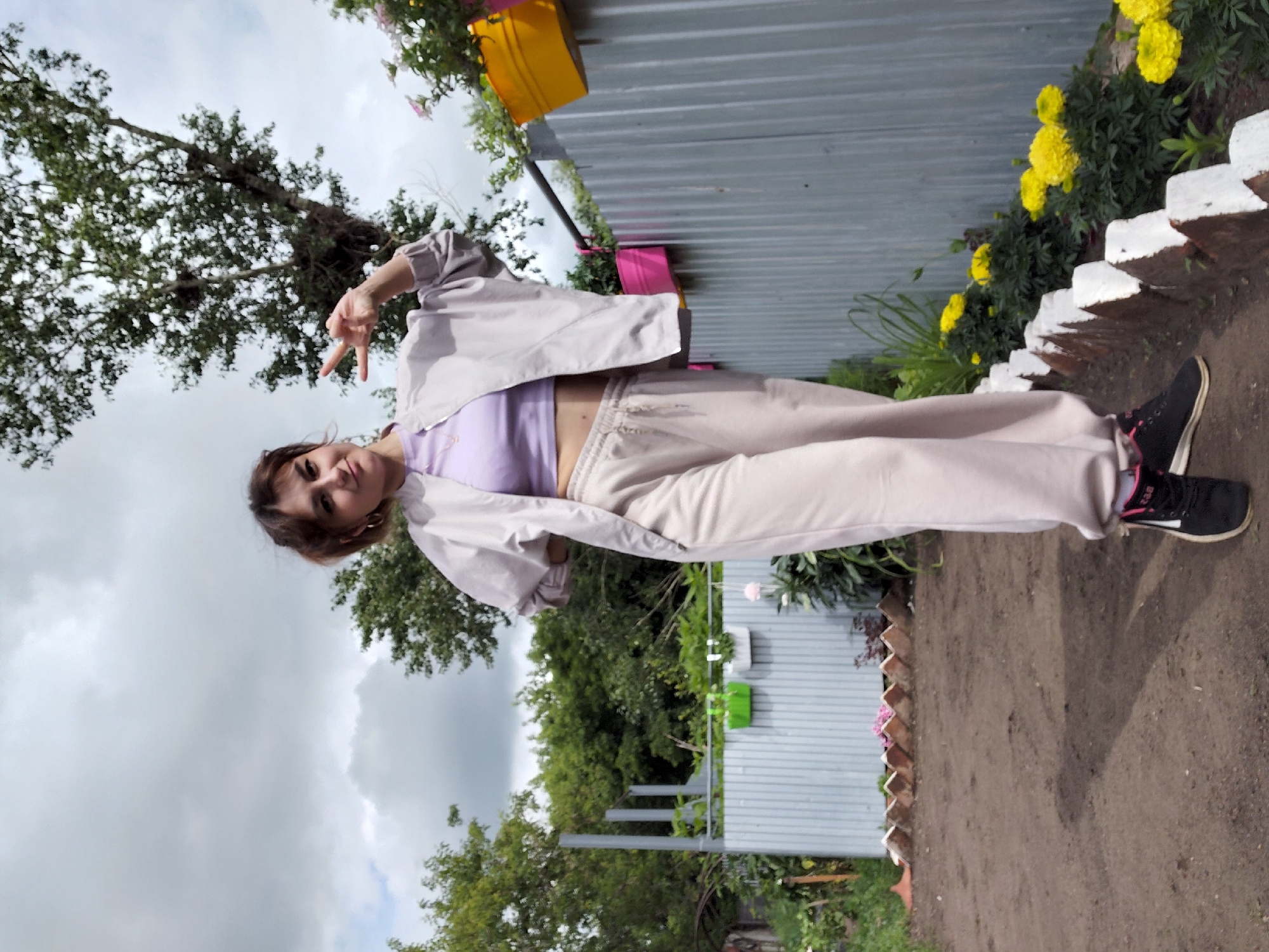 A young woman stands in a dirt yard, posing for a photo. She is wearing a pale lavender crop top, an oversized beige jacket, and matching beige sweatpants. Her hair is pulled back in a messy bun, and she is wearing hoop earrings and black sneakers with pink accents. She is making a peace sign with her left hand and resting her right hand on her hip. Behind her is a corrugated metal fence, with several potted plants hanging on it, including a bright yellow pot and a bright pink pot. To the left of the woman are more trees and foliage. To the right of the woman, along the fence, are rows of yellow marigolds. The ground is dirt and gravel, and there is a decorative border of bricks painted white along the flowerbed. The sky is cloudy and overcast. The location is identified as Tashkazgan, Russia.
