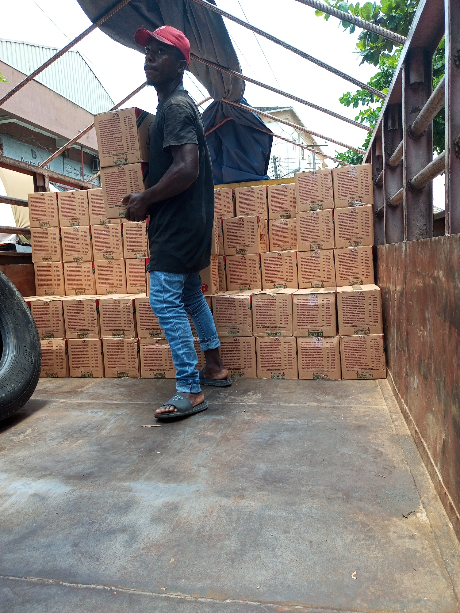 A man is depicted inside the bed of a truck, handling two stacked cardboard boxes. He has dark skin, wears a black t-shirt, light blue jeans rolled up at the ankles, and black slide sandals. A red baseball cap covers his head, and his gaze is directed upwards and to the right, showing a focused expression.
The truck bed is largely filled with numerous identical brown cardboard boxes, stacked in organized rows along the back and side, forming a solid wall of cargo. Each box displays a "JAFC" logo, columns of fine print which likely detail product information, and international handling symbols. Visible text on some boxes indicates "10 x 1.2KG". The truck floor is a dark, worn metal surface. A large black tire is partially visible on the lower-left side of the frame.
Above the man and cargo, metal rods and ropes, part of the truck's structure, are visible, along with a dark tarpaulin covering some goods. Beyond the truck, a bright, overcast sky dominates the background. Portions of buildings and green tree foliage are also visible, including a building with a sign that partially reads "Auto P..." or "Auto Del...". The scene suggests a commercial activity, such as loading or unloading goods, taking place in an urban environment like Satellite Town, Nigeria, during daylight hours.