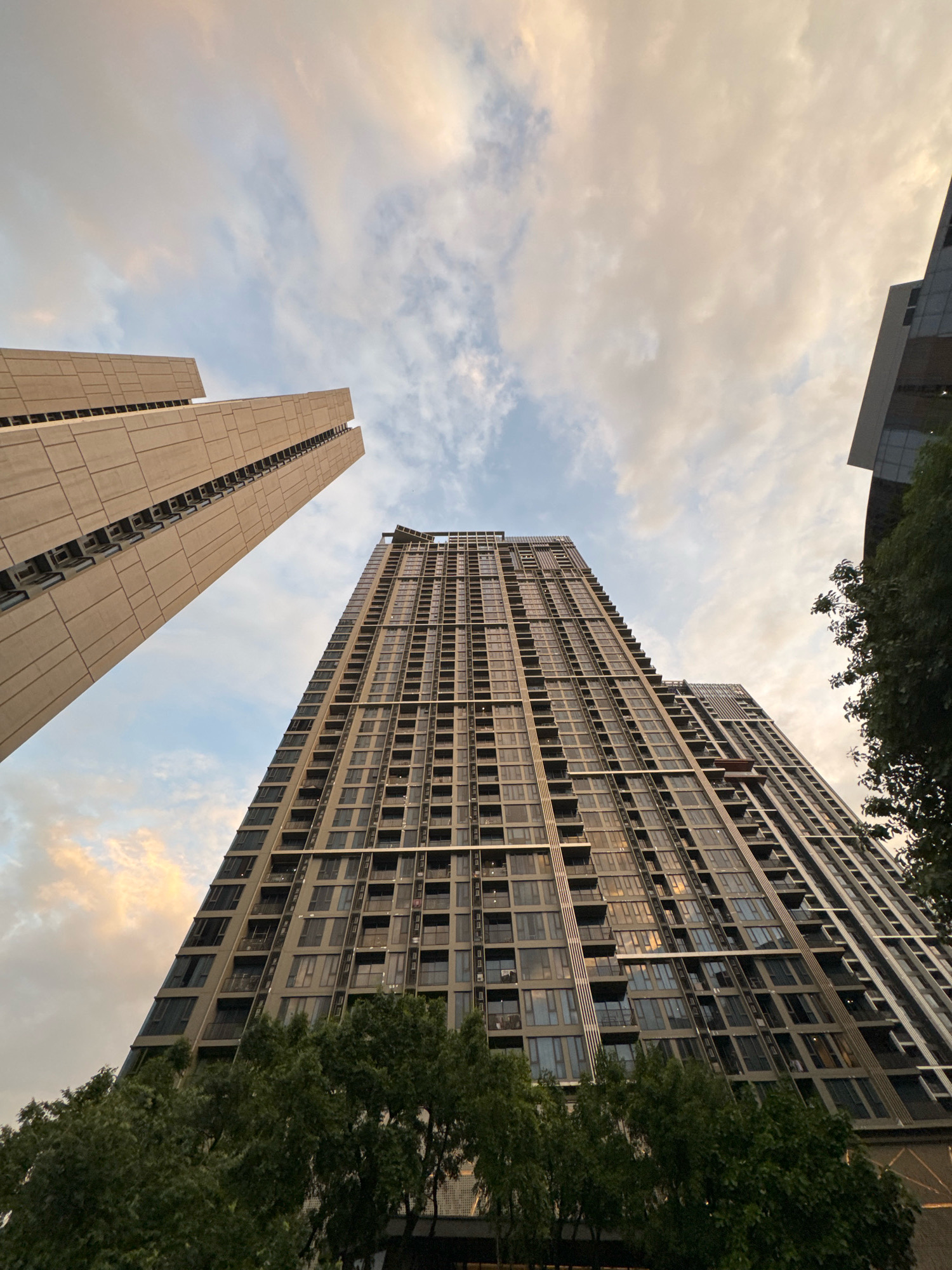 This low-angle shot captures the imposing modern architecture of several high-rise buildings against a dusky sky in Bangkok, Thailand. In the foreground, lush green trees partially obscure the lower levels of two prominent skyscrapers. The taller, central building is a dark, glass-fronted structure with countless windows and balconies, its height emphasized by the upward perspective. To its left, a lighter-colored, more angular building features a distinct, textured facade with rows of windows. The sky above is a canvas of soft clouds, tinged with the warm hues of sunset, creating a dramatic contrast with the urban structures. The image conveys a sense of urban scale and the interplay between nature and man-made design in a bustling city.