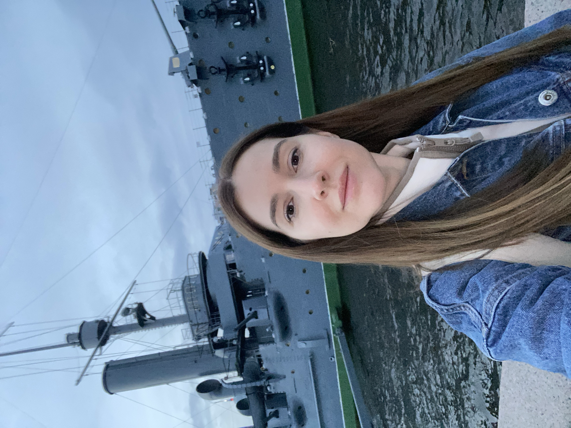 A young woman with long brown hair and a denim jacket is taking a selfie. She is smiling slightly, looking towards the camera. In the background, the stern of a large, grey ship is visible. The ship appears to be a museum or historical vessel, possibly a cruiser, with various structures, masts, and railings. The ship is docked in water, and there is a green stripe along its hull. The sky is overcast, suggesting it might be a cloudy day, possibly late afternoon or early evening, given the subdued light. The overall scene suggests a visit to a waterfront attraction or a historical site in Saint Petersburg, Russia.