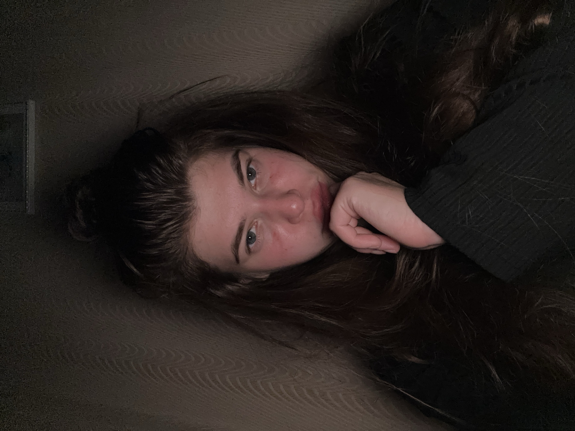 A young woman with long, dark brown hair lies on a brown, patterned surface, possibly a sofa or bed. Her hair is partially up in a bun. She is wearing a dark-colored, possibly black, ribbed sweater. Her head is tilted, and she is looking directly at the camera, resting her cheek on her hand. The lighting is dim. A portion of what appears to be a window or panel is visible in the upper left corner. There is no evidence in the image to determine its location in Мещёры, Russia.