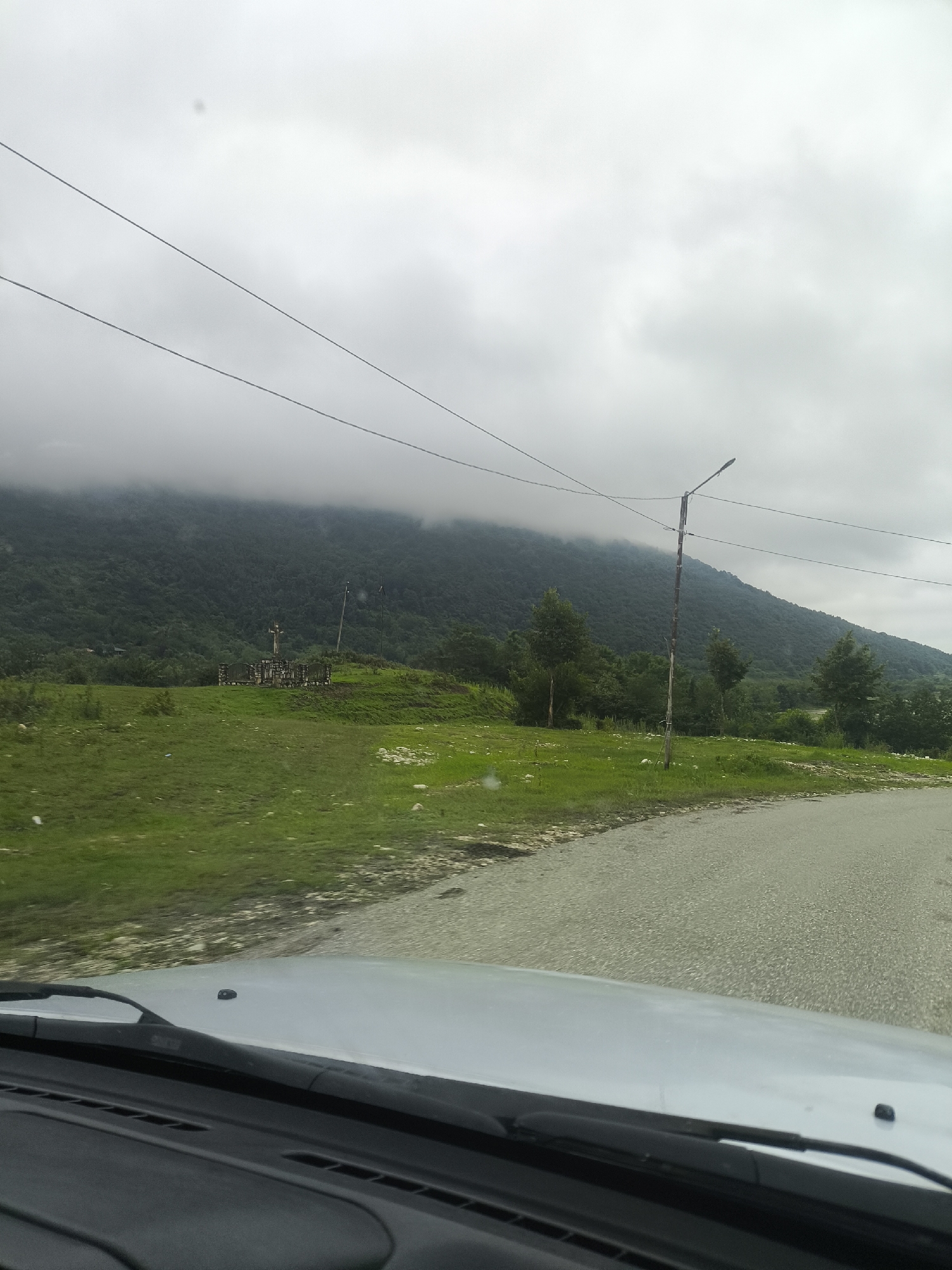 The image depicts a landscape scene from inside a vehicle, looking out towards a mountain range under a cloudy sky. In the foreground, the dashboard and windshield wipers of the car are visible. The road curves to the right, with a gray, asphalt surface. To the left of the road, a grassy hill rises, dotted with some rocks. In the mid-ground, a wooden cross monument stands on the hill, surrounded by a low stone wall. Further back, a dense forest covers the lower slopes of the mountain. The upper portion of the mountain is shrouded in thick, gray clouds, obscuring its peak. A single lamppost stands near the road on the right side of the image. The overall atmosphere is overcast and somewhat somber. The image appears to be taken in the vicinity of Skurdi, Georgia, given the characteristic mountainous terrain and lush vegetation.