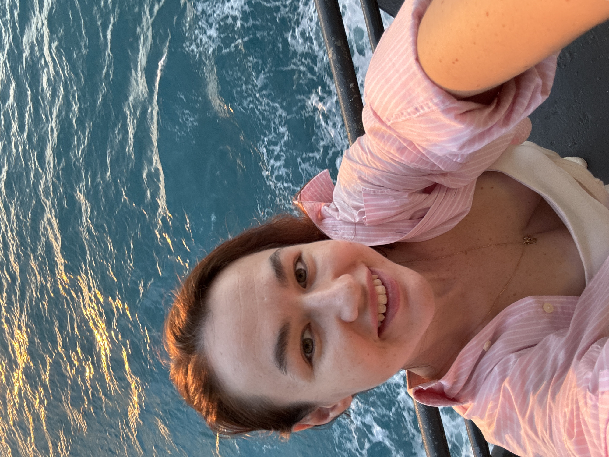 An outdoor, waist-up, selfie-style shot features a young woman with auburn hair and light skin with freckles posing in front of a body of water. She is looking directly into the camera and smiling, with her mouth open to reveal her teeth. She is wearing a light pink collared shirt with white pinstripes and a white camisole or dress underneath. A delicate gold necklace with a small pendant is visible around her neck. The water behind her appears to be the sea or an ocean, with ripples and sunlight glinting on the surface. A dark railing or barrier is visible in the foreground, partially obscuring the lower portion of the frame. The lighting suggests it may be either sunrise or sunset, with a warm glow casting over the scene. The image does not contain any identifiable landmarks or specific details that would indicate a location within the city of Unknown, Thailand.