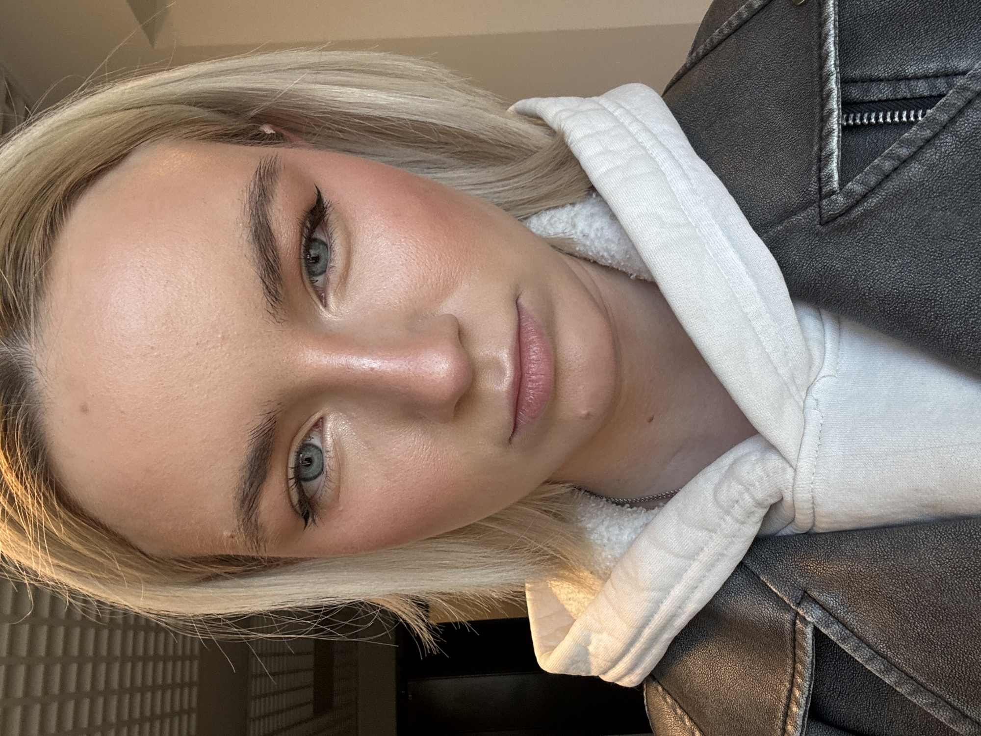 A close-up, rotated portrait captures a blonde woman. She wears winged eyeliner and pink blush. Her lips appear glossy. A white hoodie with a fleece lining is visible. A black leather jacket overlays the hoodie. The background shows a light-colored, textured wall and a dark area, possibly a doorway. The location in the city of Unknown, Russia is not discernible.