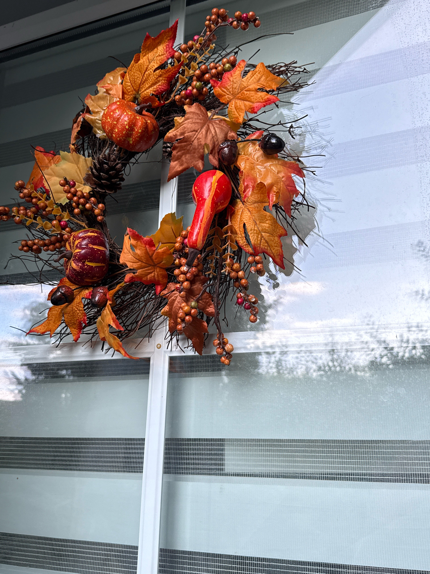 A close-up, slightly elevated, full-frame, eye-level shot shows a rustic autumn wreath hanging on a window. The wreath is composed of twigs, artificial fall leaves in shades of orange and brown, small red berries, miniature pumpkins, pinecones, and acorns. One noticeable red, gourd-shaped ornament is also attached. The wreath is positioned in the upper left quadrant of the image, with its lower edge extending towards the center. The background consists of a window with a modern roller blind that has alternating horizontal bands of sheer and opaque material. Sunlight is casting dappled shadows from the wreath and its surroundings onto the blind. The environment appears to be indoors or a sheltered outdoor space with a clear view of the sky and possibly trees through the window. There are no people or animals visible in the image. The scene is static and devoid of action. The image was captured on a bright, possibly overcast day, given the soft light and visible shadows.