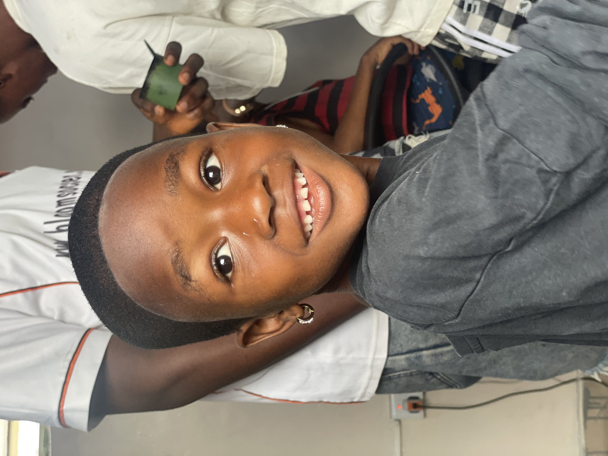 A close-up, slightly upward-angled shot captures a smiling young person with dark skin, short dark hair, and gold hoop earrings, situated indoors, possibly in Ado, Nigeria. The individual wears a gray t-shirt. To the left, a person in a white t-shirt with orange trim is partially visible. Another person is seen holding a small, green object. In the background are other individuals and items of clothing. A power outlet is visible at the lower edge of the frame.