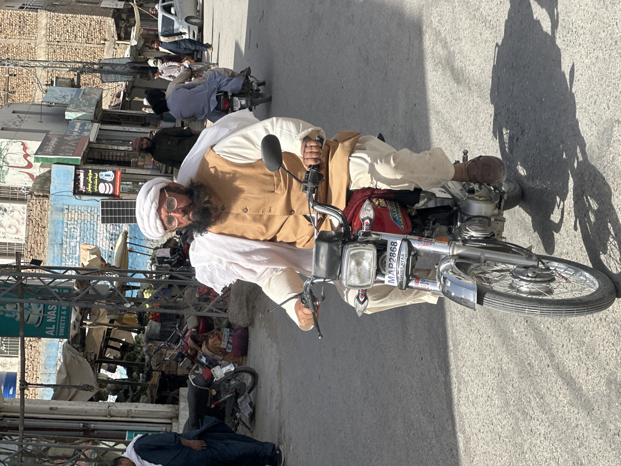 The image presents a vibrant street scene in Pishin, Pakistan, captured during bright daylight. The central figure is an elderly man with a long, dark beard and spectacles, wearing a white turban and a light brown waistcoat over a white traditional shalwar kameez. He is seated on a red motorcycle, with his hands gripping the chrome handlebars, and looks directly towards the camera with a neutral expression. The motorcycle's license plate, "MAP2868 SHINE STAR," is clearly visible.
The setting is a bustling commercial thoroughfare with a paved road and busy sidewalks. The background features various buildings, some with exposed brickwork and others with blue and white painted surfaces, typical of Pakistani architecture. Several other men, also dressed in traditional attire, are scattered throughout the scene, either walking, standing, or riding motorcycles, adding to the dynamic atmosphere of a market or town center. On the lower left, a man in a dark blue garment is visible, appearing to sit or rest on the ground.
The street is characterized by its commercial activity and various elements. Metal scaffolding or utility poles are prominent, and numerous commercial signs adorn the storefronts. One sign prominently displays "AL NAS SWEETS & B" (likely "Sweets & Bakers"), with accompanying Urdu text identifying it as "سويتس بيكرز اینڈ ملک شاپ" (Sweets Bakers and Milk Shop). A blue water barrel marked "500" is also visible. Strong, defined shadows cast by people and objects, including a large shadow of the motorcycle and rider on the road, indicate a sunny day, likely around midday or early afternoon.