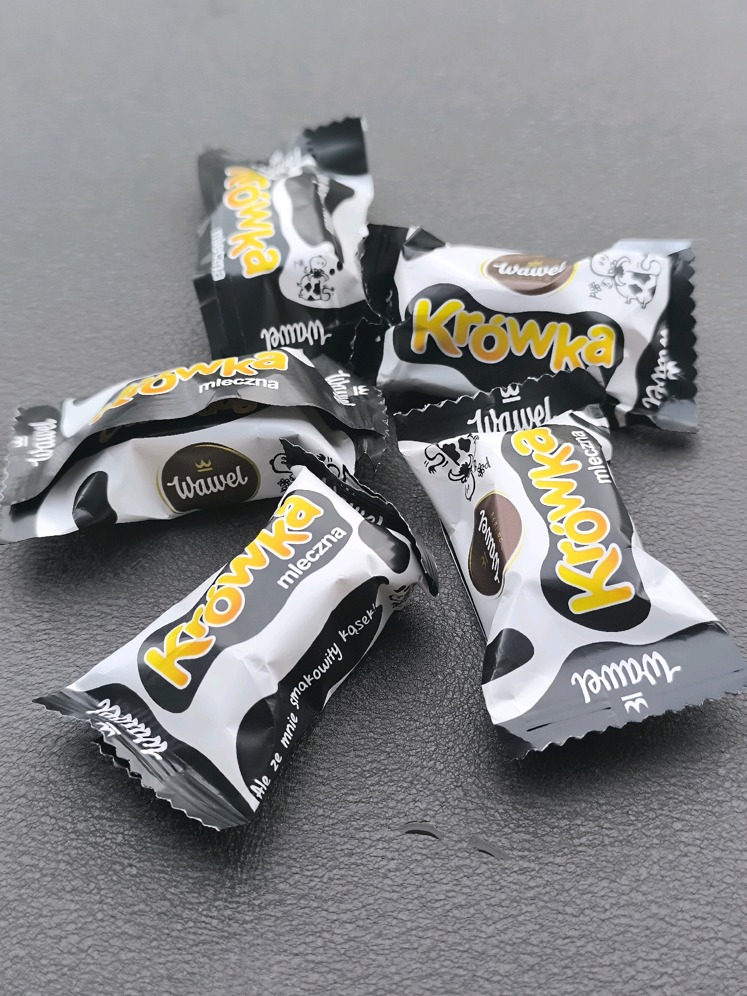 The image shows four individually wrapped candies on a dark, textured surface. The wrappers are primarily black and white with a cow-print pattern. Each wrapper features the word "Krowka" in large yellow letters with orange outlines, and "mleczna" in smaller white text. The brand name "Wawel" is also visible on the wrappers in white cursive font, sometimes within an oval. One wrapper has a small drawing of a cow and the text "Ale ze mnie smakowity krasek!". The candies are arranged in a scattered, somewhat overlapping manner. The surface appears to be artificial, possibly a tabletop or a mat. There is no indication of any specific location in Dercekliai, Lithuania.