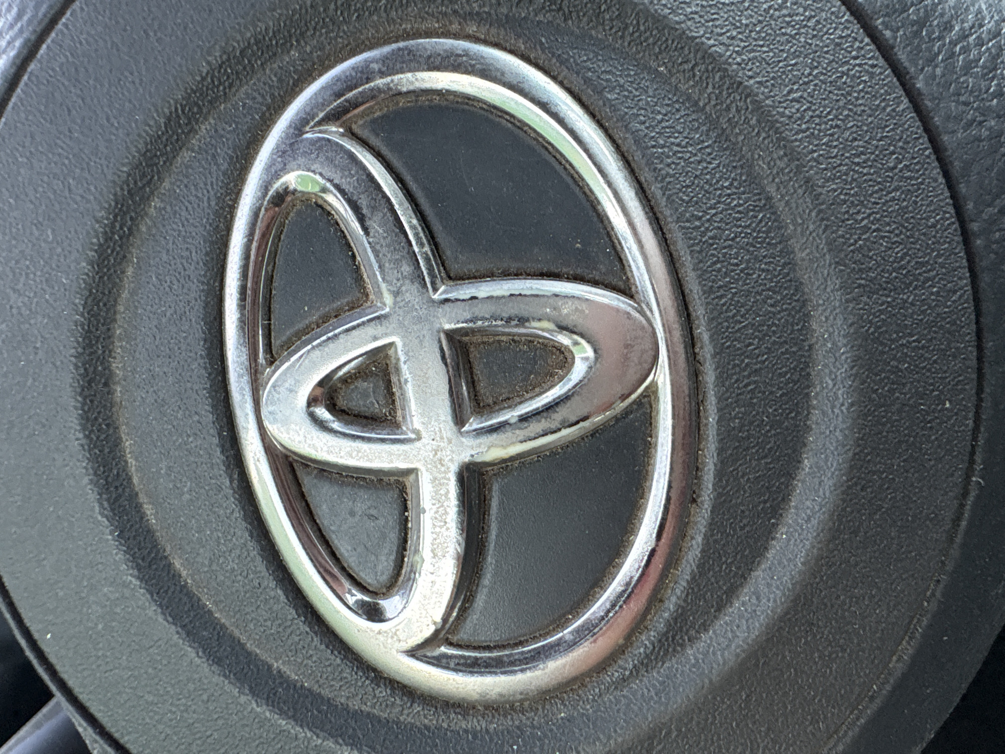 A close-up, eye-level shot shows the Toyota logo on a black steering wheel. The metallic silver logo, consisting of three interlocking ovals, is centered and slightly raised against a dark, textured background. The steering wheel surface appears to be made of a coarse plastic or leather material. The lighting is even, highlighting the shine of the logo and the matte finish of the steering wheel. The image is cropped tightly, focusing exclusively on the emblem and the immediate surrounding area of the steering wheel. There is no indication of the environment beyond the steering wheel, nor are there any people present.