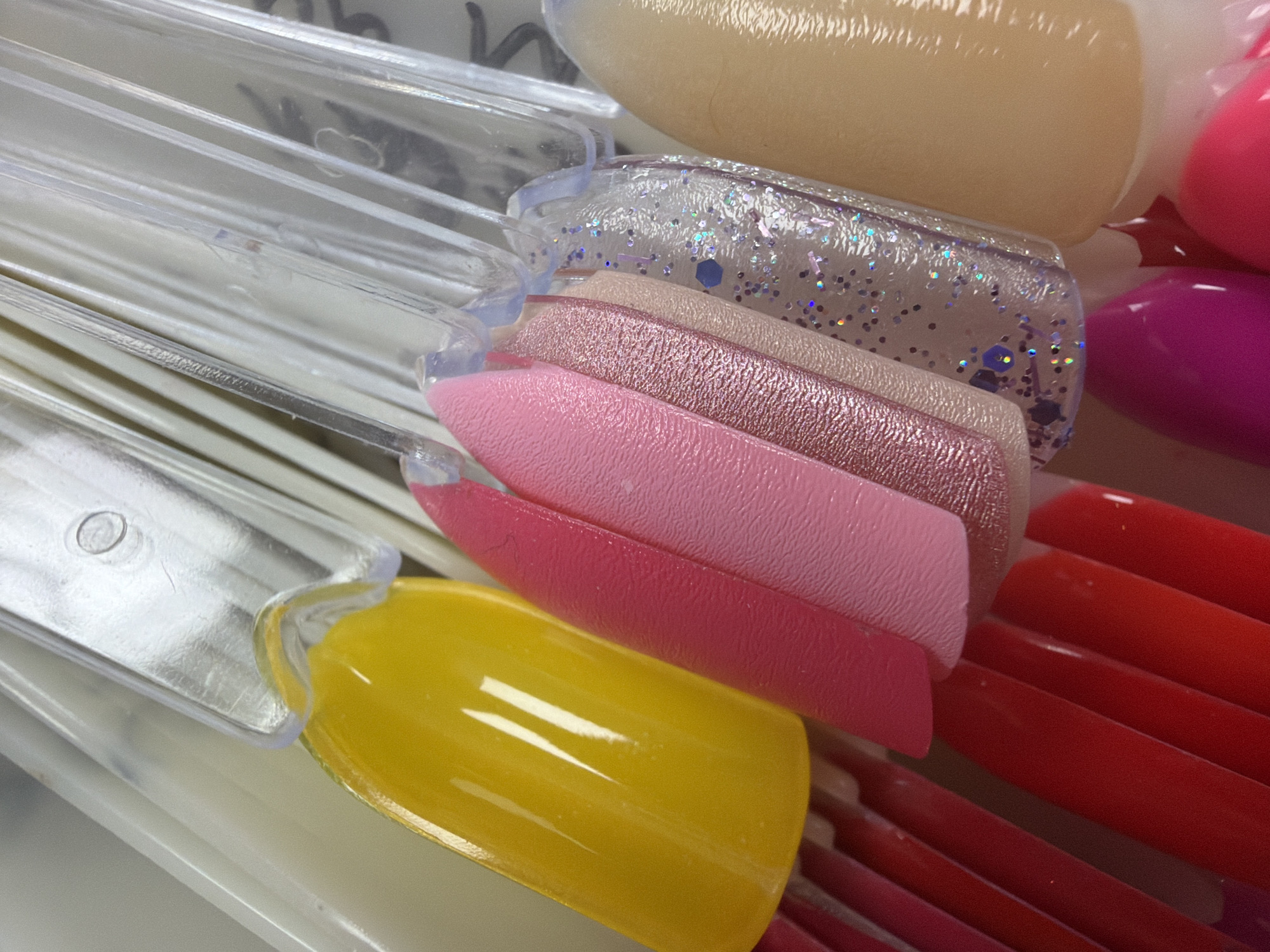 The image shows a close-up, high-angle shot of a collection of artificial nail tips. The nail tips are arranged in a fan-like formation and are in various colors and finishes, including yellow, several shades of pink, a glittery silver, a light beige, and at the bottom, bright pink and magenta. The tips are mounted on clear plastic holders, which are attached to a white backing. The lighting is bright and appears to be from above, creating reflections on the glossy surfaces of the nail tips. There are no people or recognizable landmarks present. The image does not contain any information that would allow for its location to be identified in Budva, Montenegro, or any other specific geographical location.