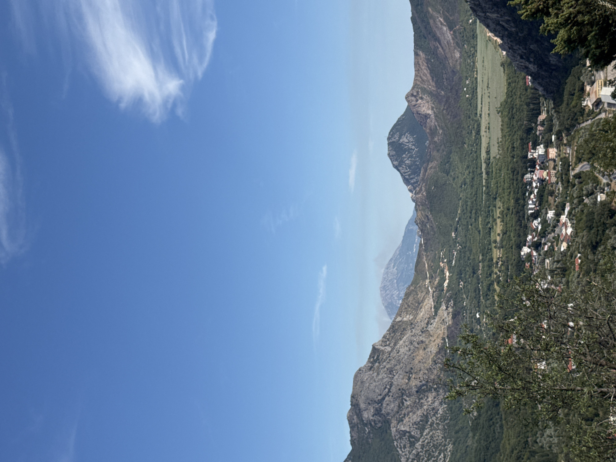 A high-angle, full outdoor shot captures a mountainous landscape under a clear blue sky with scattered wispy clouds. In the foreground, a leafy green tree partially obscures a vista of rolling, verdant hills and rugged, rocky mountains. A small village with red-roofed buildings is nestled in the valley below, surrounded by lush vegetation and winding roads. In the background, more distant mountains rise, with one peak appearing particularly prominent. The scene suggests a rural setting in Kaluđerac, Montenegro, characterized by its natural beauty and a sense of tranquility.