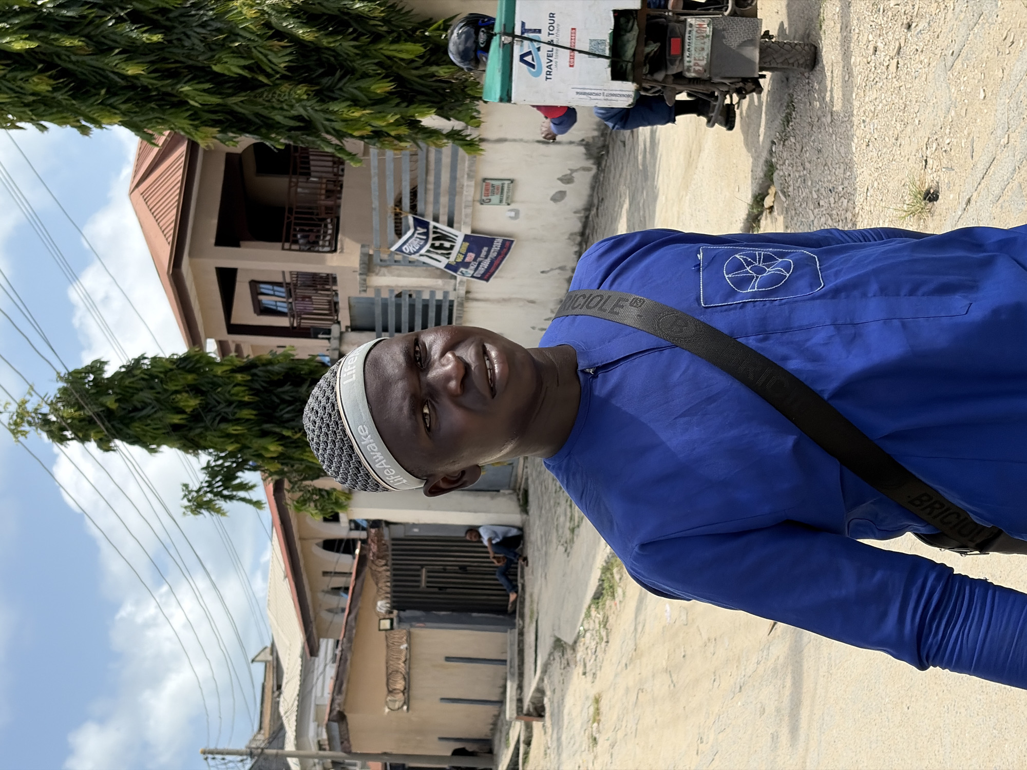 A man wearing a blue shirt, a skull cap with the word "LifeAwake" on the band, and a black strap across his torso stands on a street in Igboefon, Nigeria. The shirt has a stitched emblem on the upper left. In the background, there is a building with a brown roof and a gray metal fence. A sign advertising "PRATY LUXURY" and "IKENY" is visible. A motorcycle with a "TRAVEL & TOUR" container strapped to the back is also present. The ground appears to be a mix of concrete and dirt. There is a man in a white shirt sitting inside of the metal fence gate. Trees and overhead wires are visible.