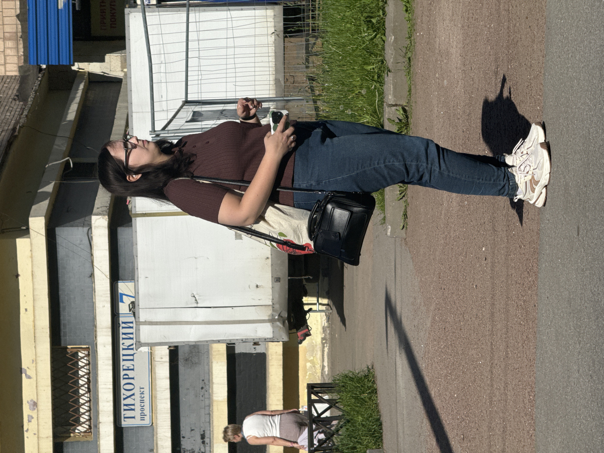 The image captures a candid street scene on a bright, sunny day in what is identified as Russia. The primary subject is a woman with dark hair and glasses, looking upwards and slightly to her right. She is wearing a dark brown ribbed short-sleeved top, blue jeans, and white and beige chunky sneakers. She holds a light-colored smartphone with her right hand and carries a black shoulder bag over her left shoulder, with a light-colored canvas tote bag also visible. A ring is visible on her left ring finger.
The woman is standing on a brownish asphalt sidewalk next to a darker gray asphalt road. Long, distinct shadows are cast on the ground, suggesting it is either mid-morning or late afternoon. To her left, there are buildings with varying facades, some featuring yellowish lower sections and white or gray upper walls. A blue corrugated metal structure is visible on a building further back. A strip of grass with dandelions runs along the sidewalk, separated by a low fence.
In the background, another person, seen from behind, is bent over near some greenery and a black metal fence.
Visible text on a street sign clearly reads "ТИХОРЕЦКИЙ" (Tikhoretsky) and "проспект" (prospekt), along with the number "7" in a blue rectangular box, indicating "Tikhoretsky Prospekt 7". Another partially obscured sign shows the words "ПРИЯТНЫ" (Priyatny - pleasant) and "ПОКУП" (Pokup - purchase/buying), possibly part of "Приятных покупок!" (Happy shopping!).