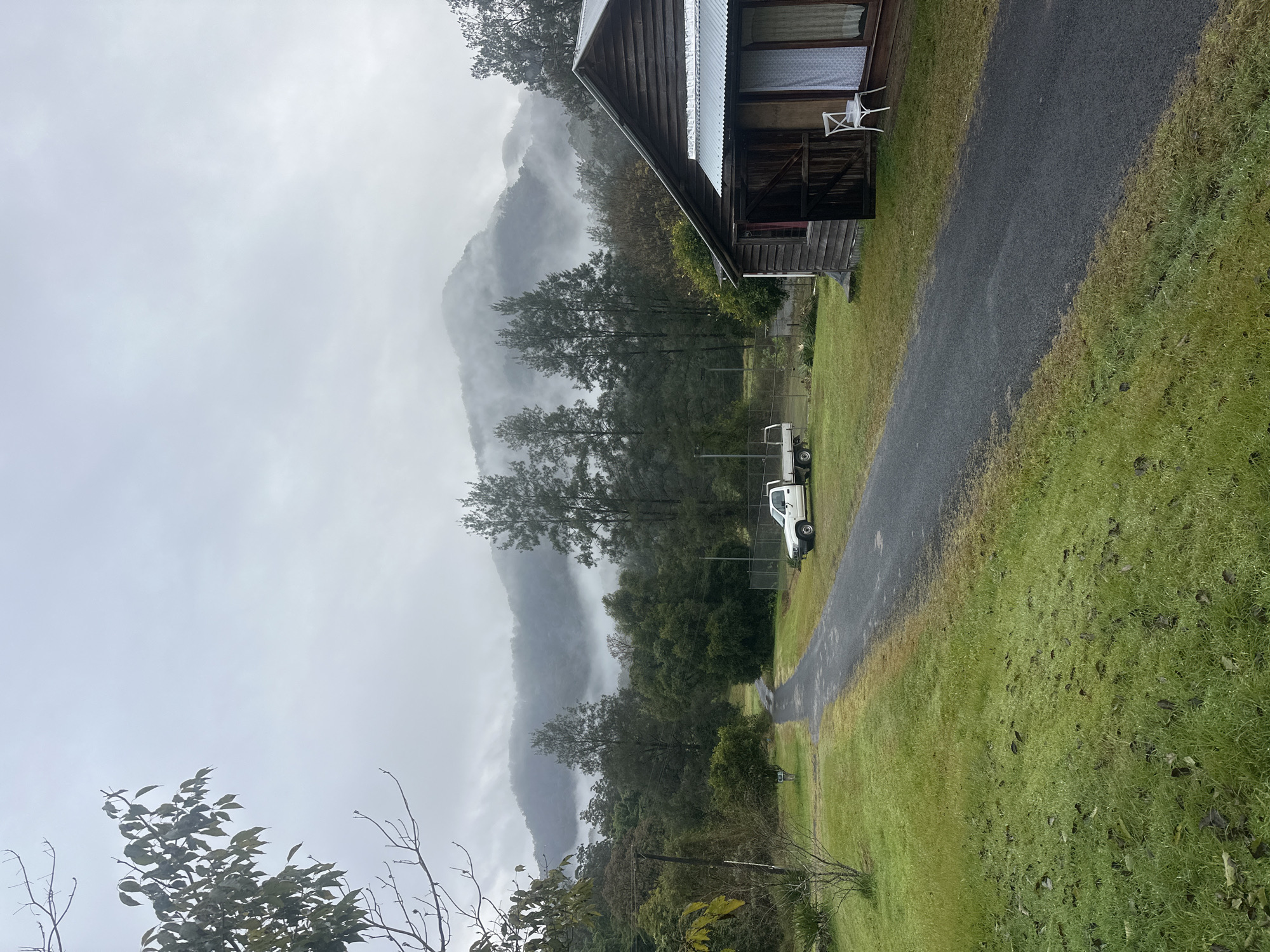 The image depicts a rural scene in Thora, Australia, under an overcast, misty sky. A dark asphalt driveway curves from the foreground into the middle ground, bordered by lush green grass. On the right side of the driveway, a rustic wooden building with a corrugated iron roof stands. The building has a covered porch with a white chair placed outside. To the left of the driveway, a white utility vehicle with a flatbed is parked. Beyond the vehicle, a fence runs along the property line. In the background, a dense forest transitions into rolling hills that are partially obscured by mist and fog, creating a soft, diffused light across the landscape. Tall, slender trees dot the middle ground, interspersed with the darker foliage of the forest. Bare branches of a tree in the upper left corner frame the top of the image. The ground appears damp, suggesting recent rain.