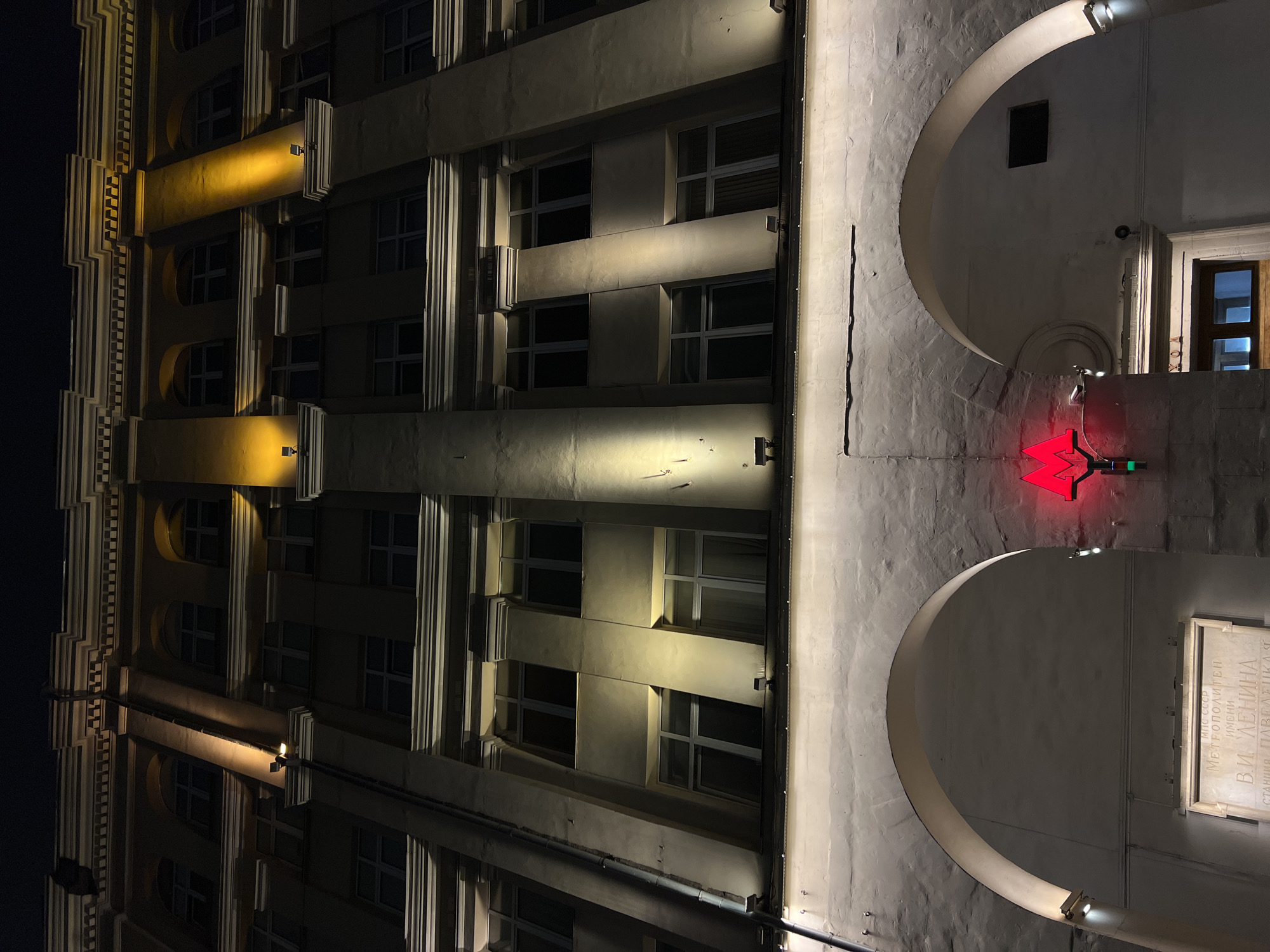 The image depicts a building at night. The left side shows an illuminated multi-story facade with windows and architectural details highlighted by warm-toned lights. The right side reveals a section of the building with an entrance marked by arches and a bright red "M" symbol, indicative of a Metro station. A plaque inscribed with Cyrillic text is also visible next to the entrance. The building appears to be in Moscow, Russia, judging from the style of architecture and the Metro symbol.
