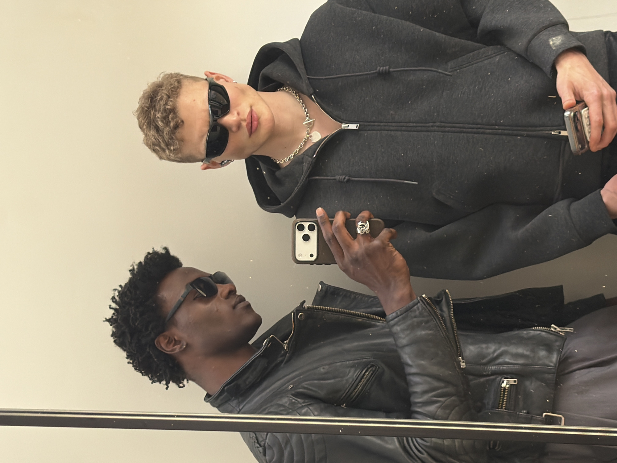 A reflection captures two individuals: one, a light-skinned person with short curly blonde hair wearing sunglasses, a silver chain necklace, and a dark gray hooded zipper sweatshirt, holding a small, possibly metallic, rectangular object. The other person, with dark skin and short dark curly hair, is wearing sunglasses and a black leather jacket, holding a smartphone. They appear to be in a public restroom with a reflective surface, possibly a mirror. The location is the city of London, United Kingdom.