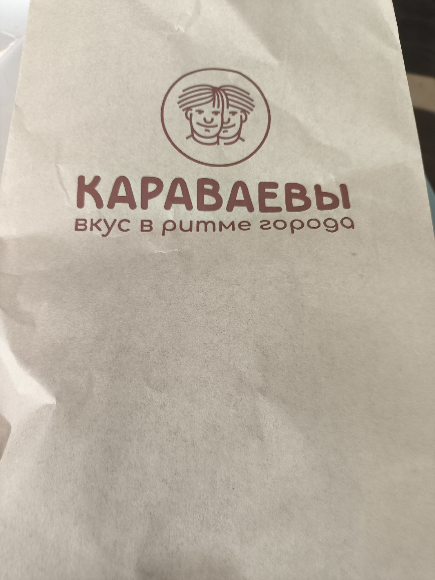 A close-up shot depicts a paper bag with the logo of a brand named "KAPABAEBbI". The logo is an oval shape containing two stylized faces and, below it, the phrase "Bkyc B pumme ropoga", both in reddish-brown. The bag is creased and appears to be made of light-colored paper.