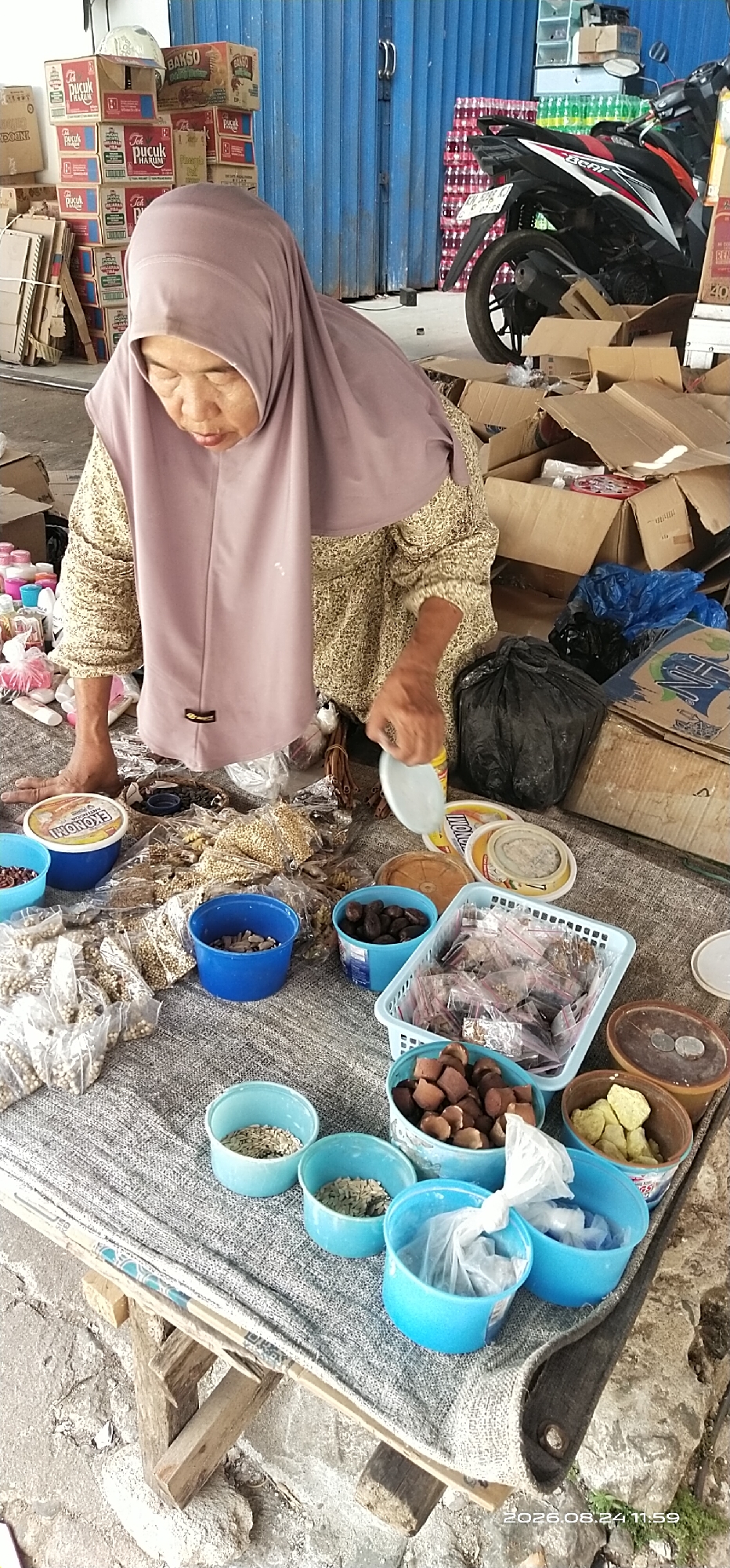 The image depicts a street market scene in Tamiang Layang, Indonesia. In the foreground, an elderly woman wearing a light purple hijab and a patterned long-sleeved shirt is bent over a display of goods. She appears to be sorting or examining small items, possibly spices or seeds, which are arranged in various containers, including blue plastic bowls and small plastic bags. Several containers of what appear to be food items or ingredients are visible, including round tubs with labels like "EKONOMI". Behind the woman, there are stacks of cardboard boxes and what looks like a motorcycle. The background reveals a blue corrugated metal roll-up door, suggesting a storefront or warehouse setting. The ground is covered with a textured mat, and the overall environment appears to be outdoors, possibly in a back alley or an open market area. The lighting indicates it is daytime.