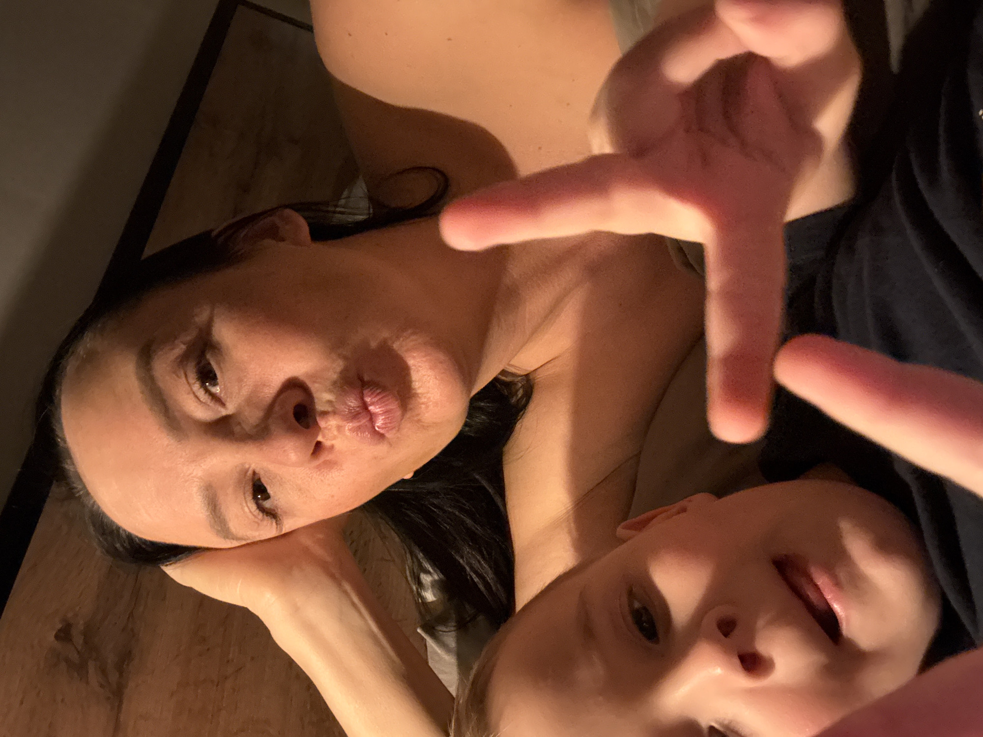 This close-up, low-light photograph, taken in Chaikovsky, Russia, captures an adult woman and a child lying down in an intimate, playful moment. The image is oriented diagonally, with warm, soft lighting illuminating their faces.
The adult woman, positioned in the upper left of the frame, has dark hair and is looking directly at the camera with a "pouty kiss" expression, her lips puckered. Her head rests on her hand, and her skin glows with the warm light.
Below her, partially visible in the lower right, is the child. The child's face is lit from the front, showing their mouth slightly open and an eye looking towards the camera. A hand, likely belonging to the child, is prominently featured in the foreground, making a "peace" sign or V-shape gesture close to the lens, creating a blurred effect. The child appears to be wearing a dark blue top.
The setting appears to be indoors, possibly on a bed or floor, with a dark, textured surface, resembling wood or a wall, visible in the background on the left. The overall activity suggests a casual, affectionate selfie or close-up shot, emphasizing connection and playfulness. No text is visible in the image.