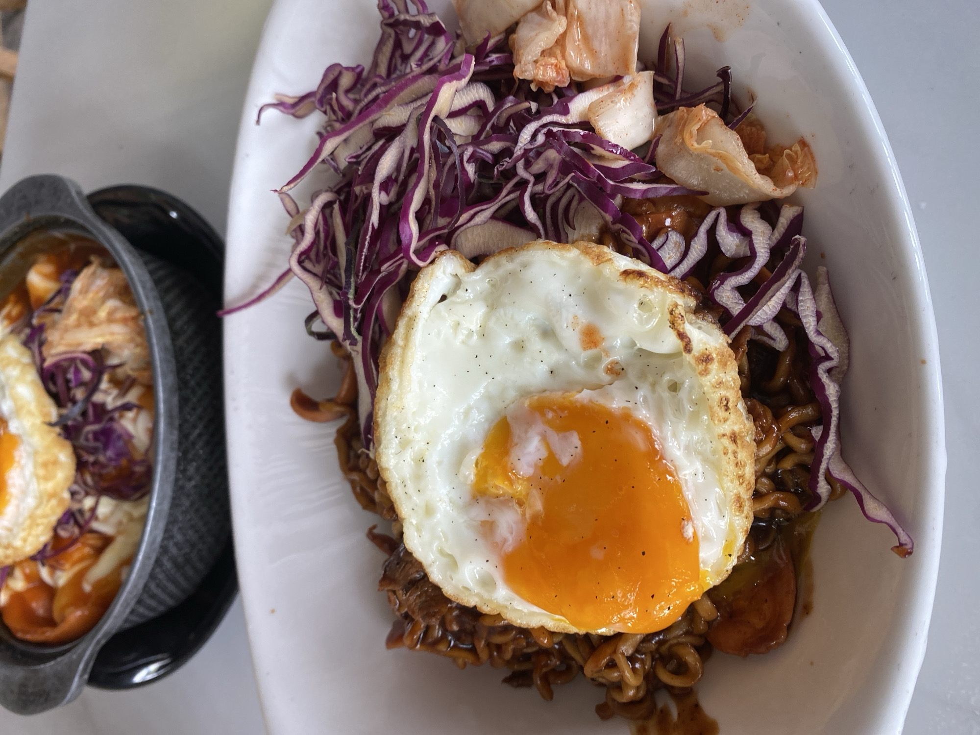 A close-up, high-angle shot shows two dishes of food on a white surface. The dish on the right, served in a white bowl, features dark noodles topped with a fried egg, shredded purple cabbage, and pale kimchi. A second dish, on the left, is served in a black, shallow pot with similar contents visible inside: noodles, a fried egg, and pieces of kimchi. The location is likely Mũi Né, Vietnam, based on the food style.